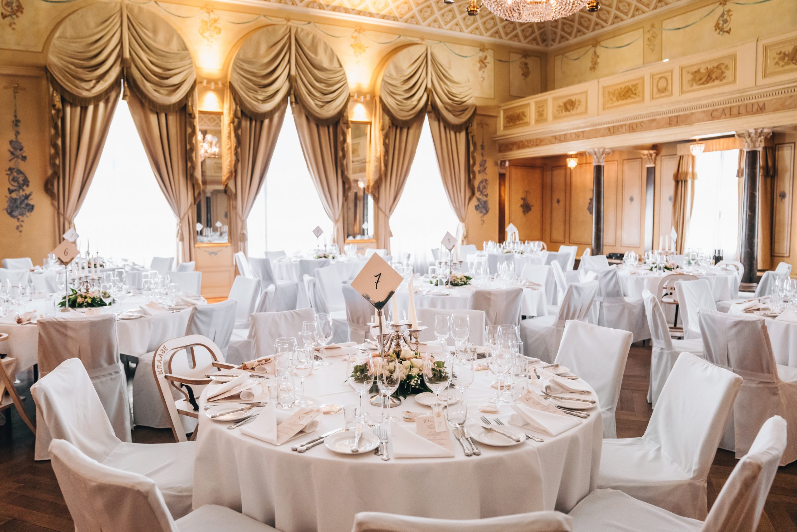 Ein formeller Bankettsaal für eine Veranstaltung mit runden, weiß gedeckten Tischen, eleganten Gedecken, blumigen Tafelaufsätzen und goldenen Vorhängen, die hohe Fenster in einem geschmückten Raum mit hohen Decken einrahmen.