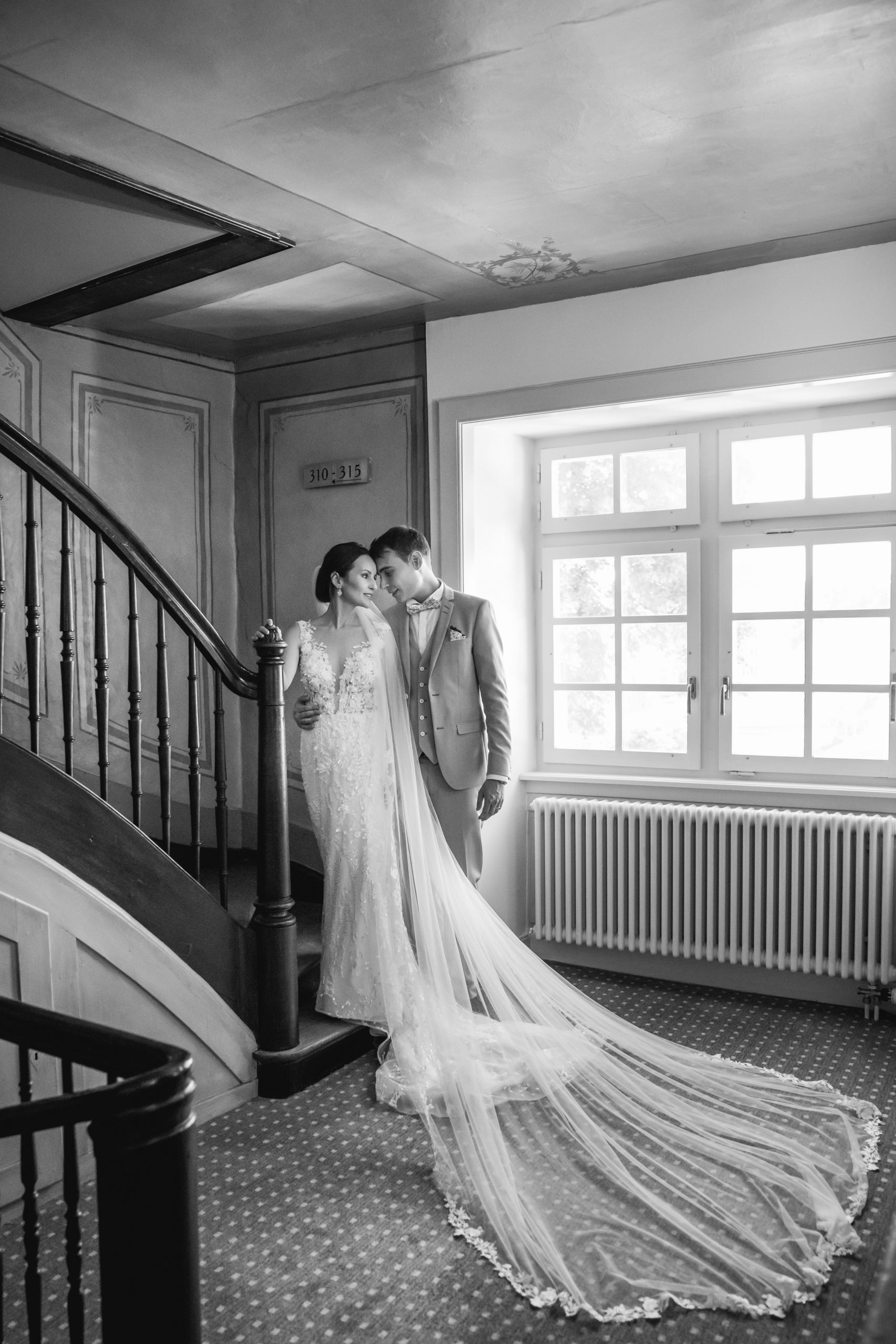 Eine Braut und ein Bräutigam stehen liebevoll zusammen an einer Treppe, die Braut in einem langen Spitzenkleid mit einem langen Schleier, der die Stufen hinunterhängt, während das Sonnenlicht durch ein Fenster neben ihnen fällt.