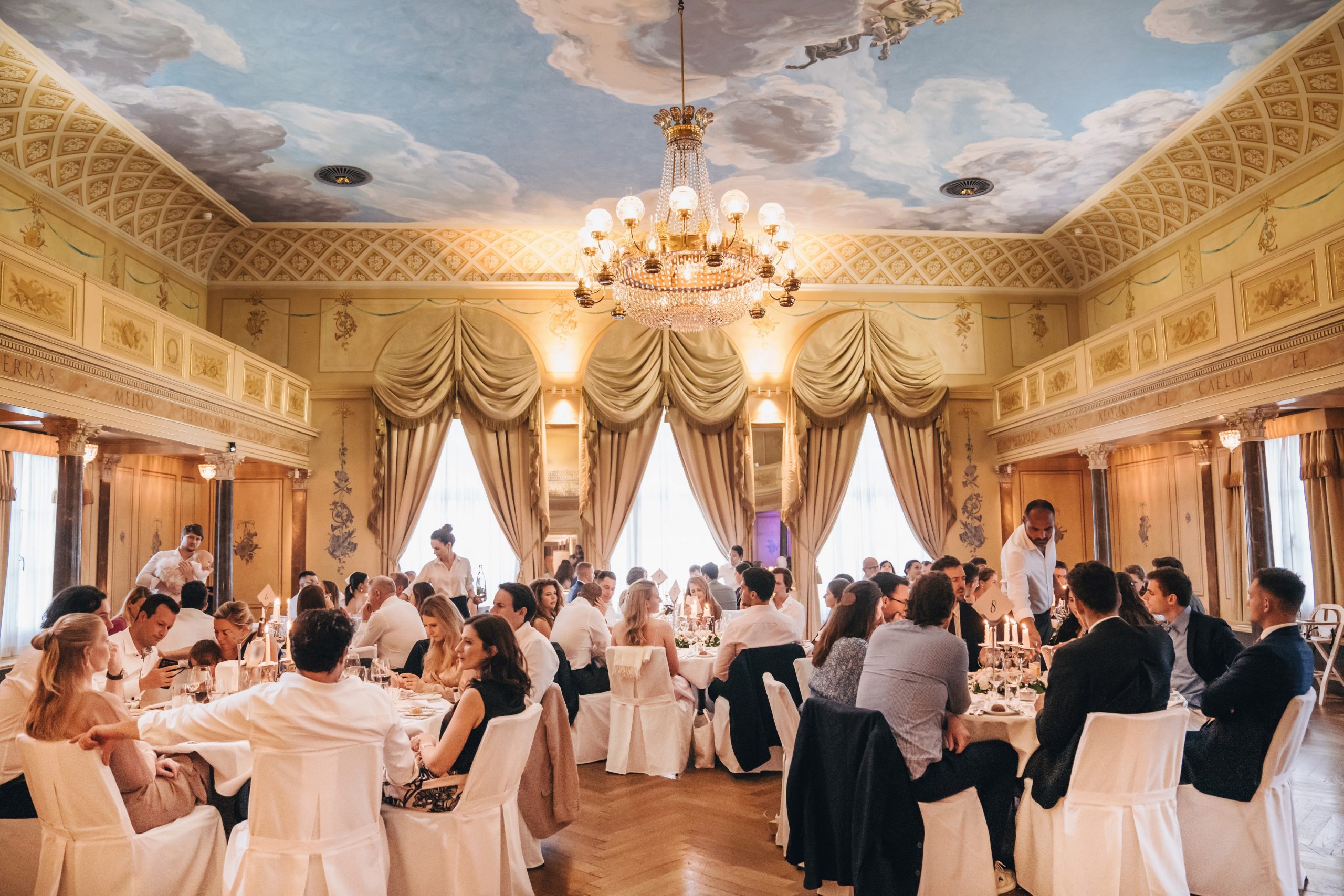 Eleganter Bankettsaal mit kunstvollem Dekor und einer bemalten Decke, gefüllt mit formell gekleideten Personen, die an runden Tischen sitzen und unter einem großen Kronleuchter speisen und sich unterhalten. Durch die hohen Fenster strömt natürliches Licht.