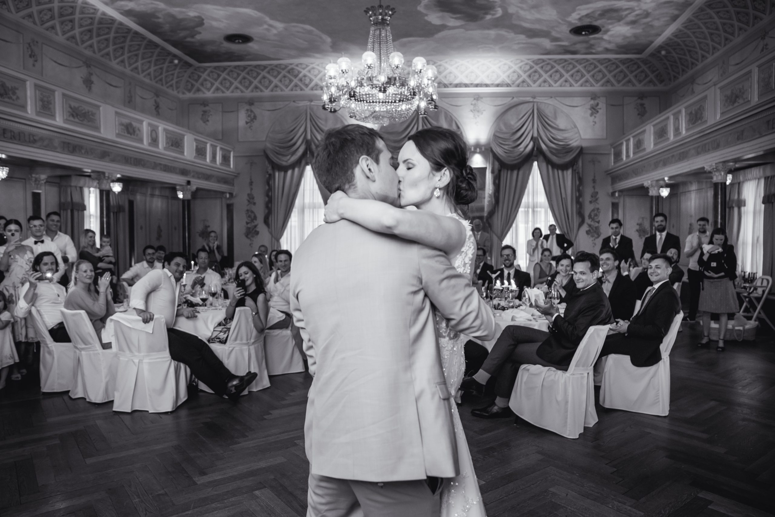 Eine Braut und ein Bräutigam tanzen ihren ersten Tanz in einem eleganten Ballsaal, umgeben von sitzenden und stehenden Gästen, die sie beobachten. Die Szene ist formell, mit Kronleuchtern und kunstvollen Dekorationen. Das Foto ist in schwarz-weiß gehalten.