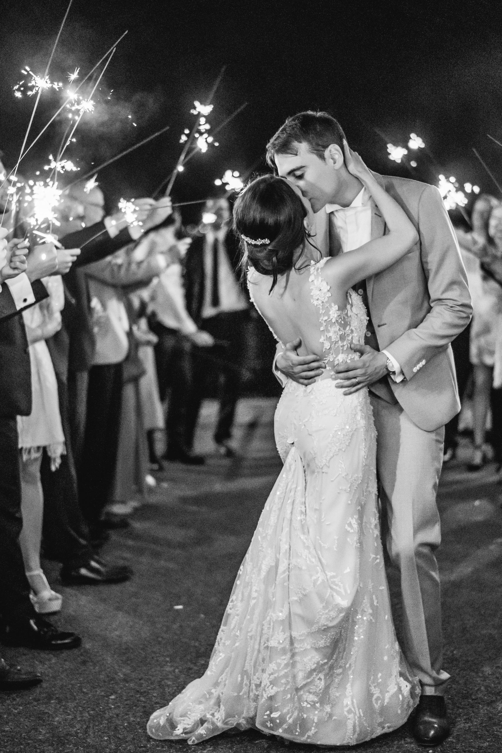 Braut und Bräutigam küssen sich bei ihrer nächtlichen Verabschiedung, umgeben von Gästen mit Wunderkerzen. Die Braut trägt ein Spitzenkleid, der Bräutigam einen hellen Anzug. Das Foto ist in schwarz-weiß gehalten.