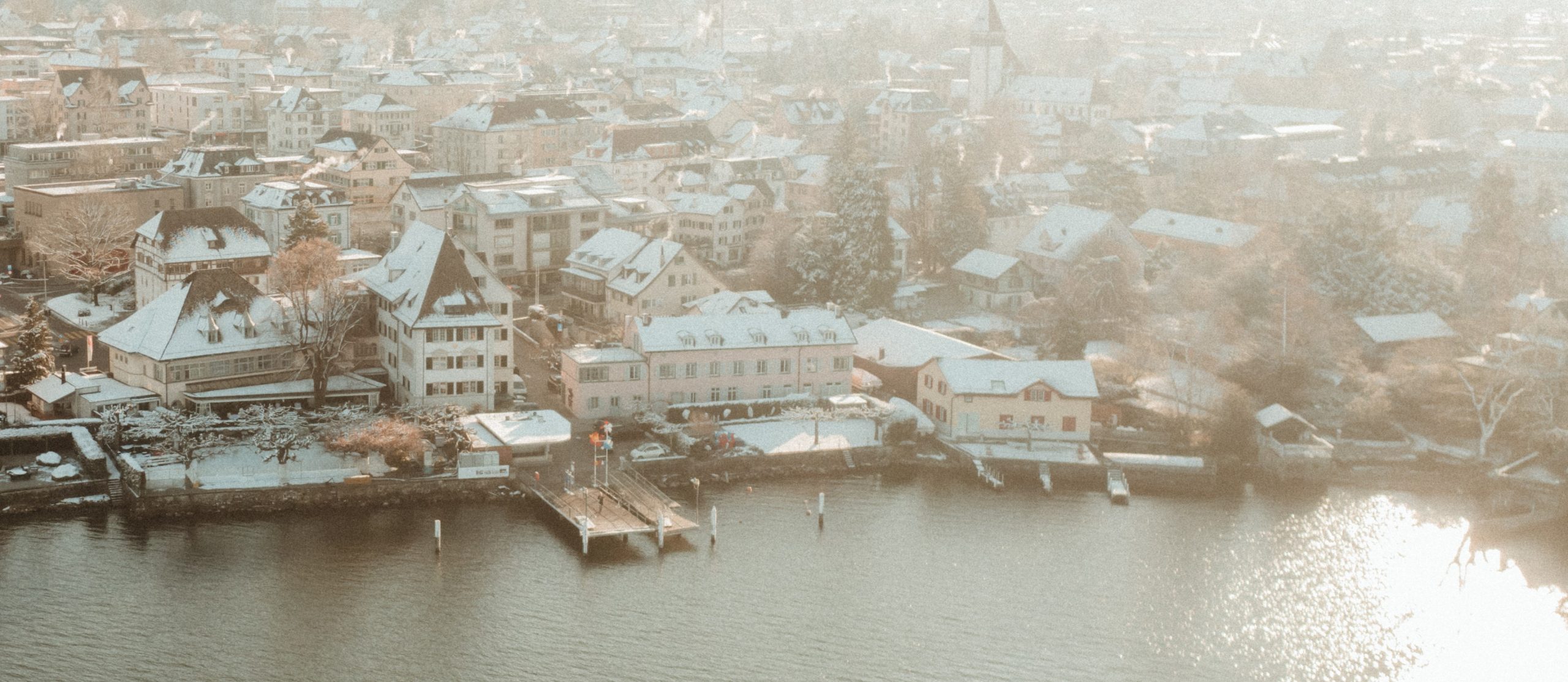 Luftaufnahme einer Stadt am Flussufer mit schneebedeckten Dächern und Gebäuden, einem kleinen Steg, der ins Wasser ragt, und Sonnenlicht, das sich auf dem Fluss spiegelt. Die Szene hat eine weiche, winterliche Atmosphäre.