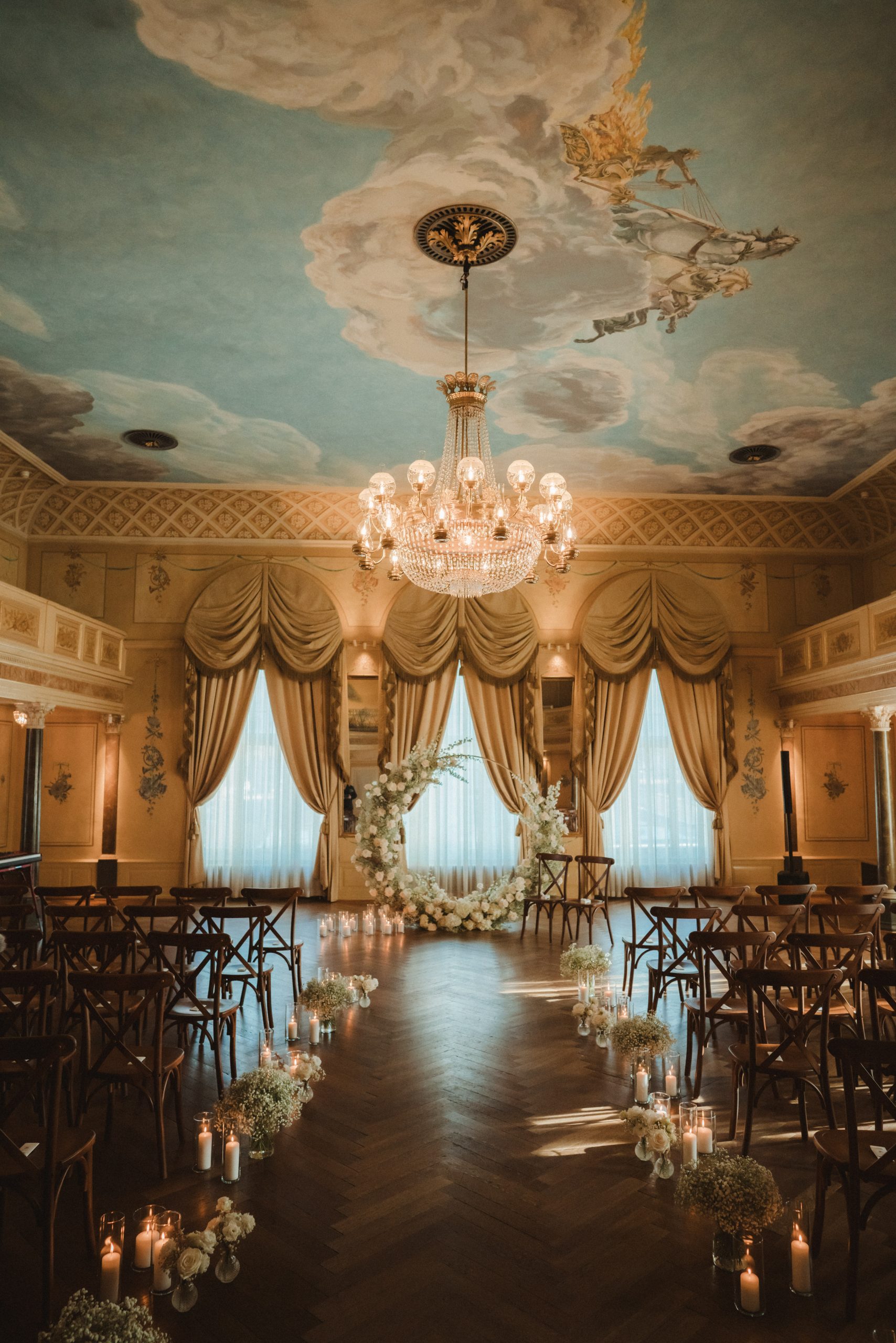 Elegante Hochzeitszeremonie im Innenbereich mit Stuhlreihen, Blumenarrangements und Kerzen in einem hölzernen Gang. Goldene Vorhänge rahmen große Fenster ein, und ein Kristalllüster hängt von einer bemalten Himmelsdecke mit Wolken und Cherubinen.
