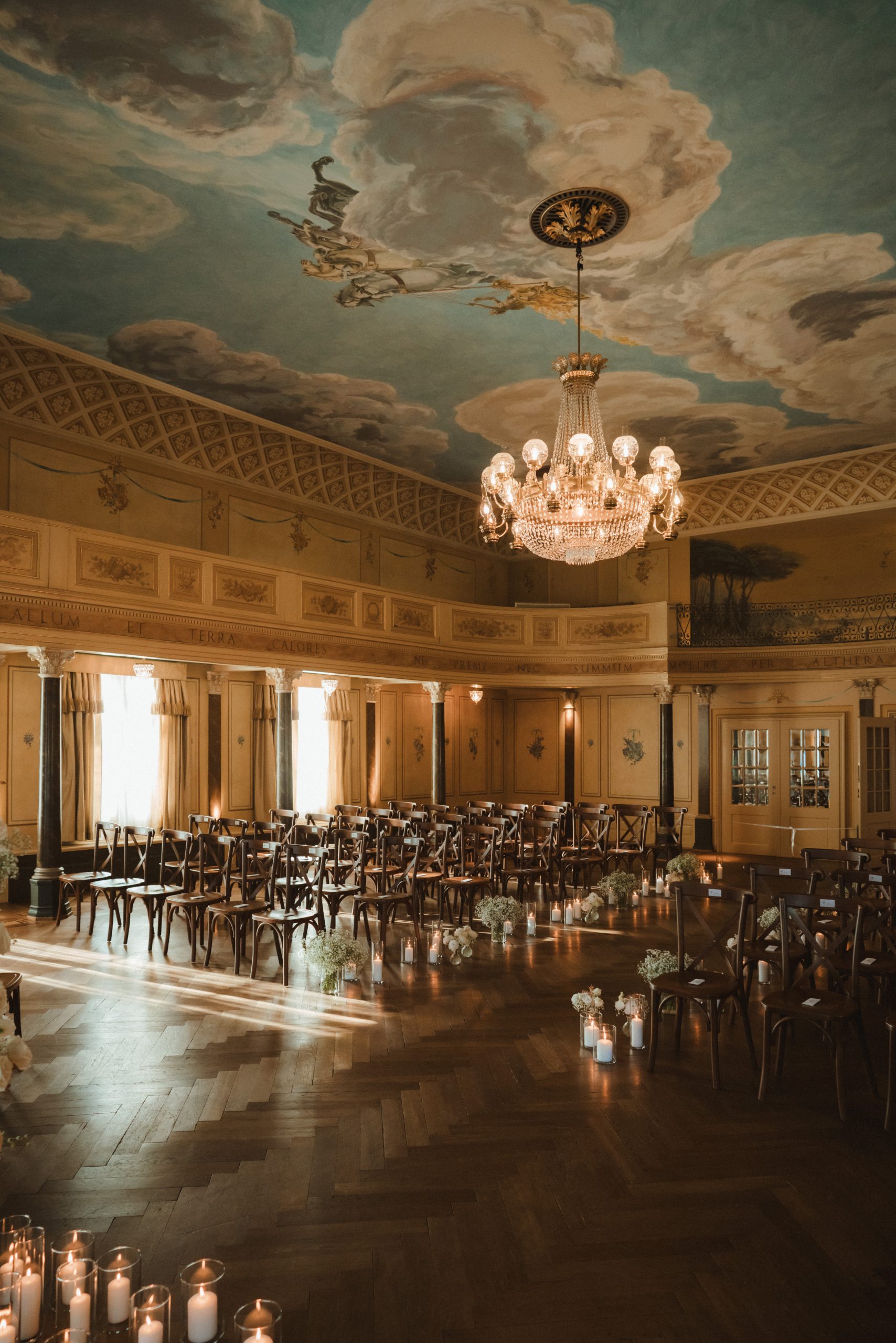 Ein großer, eleganter Raum für eine Zeremonie mit Reihen von Holzstühlen, einem mit Kerzen beleuchteten Gang, hohen Fenstern, verschnörkelten Wänden und einer bemalten Decke mit Wolken und einem Kronleuchter.