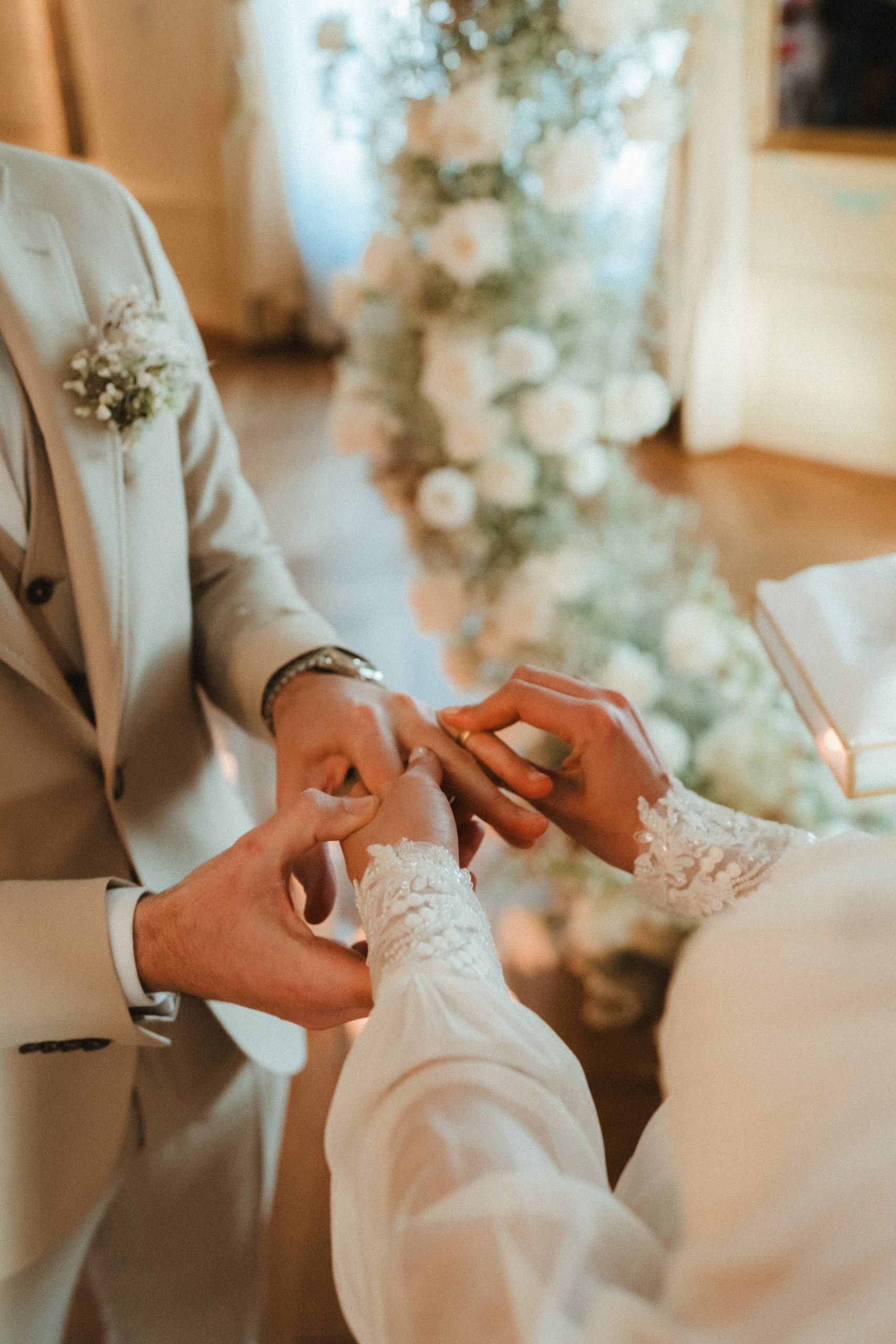 Nahaufnahme eines Paares beim Ringtausch während einer Hochzeitszeremonie, wobei die Braut dem Bräutigam einen Ring an den Finger steckt. Beide tragen helle, formelle Kleidung, und zarte weiße Blumen schmücken den Hintergrund.