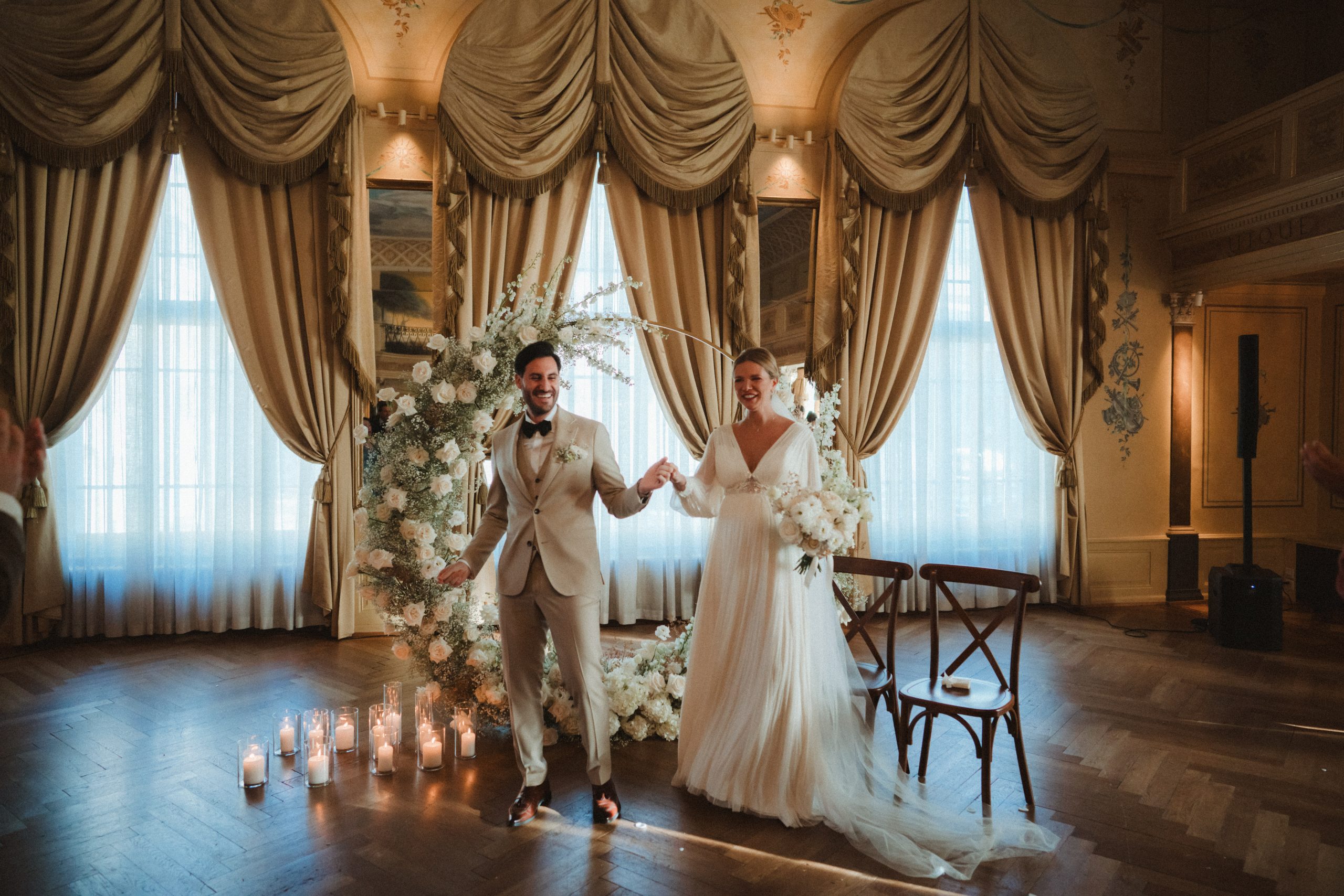 Eine Braut und ein Bräutigam stehen lächelnd und händchenhaltend in einem eleganten Raum, der mit drapierten Vorhängen, einem Blumenbogen und Kerzen geschmückt ist, und feiern ihre Hochzeitszeremonie.