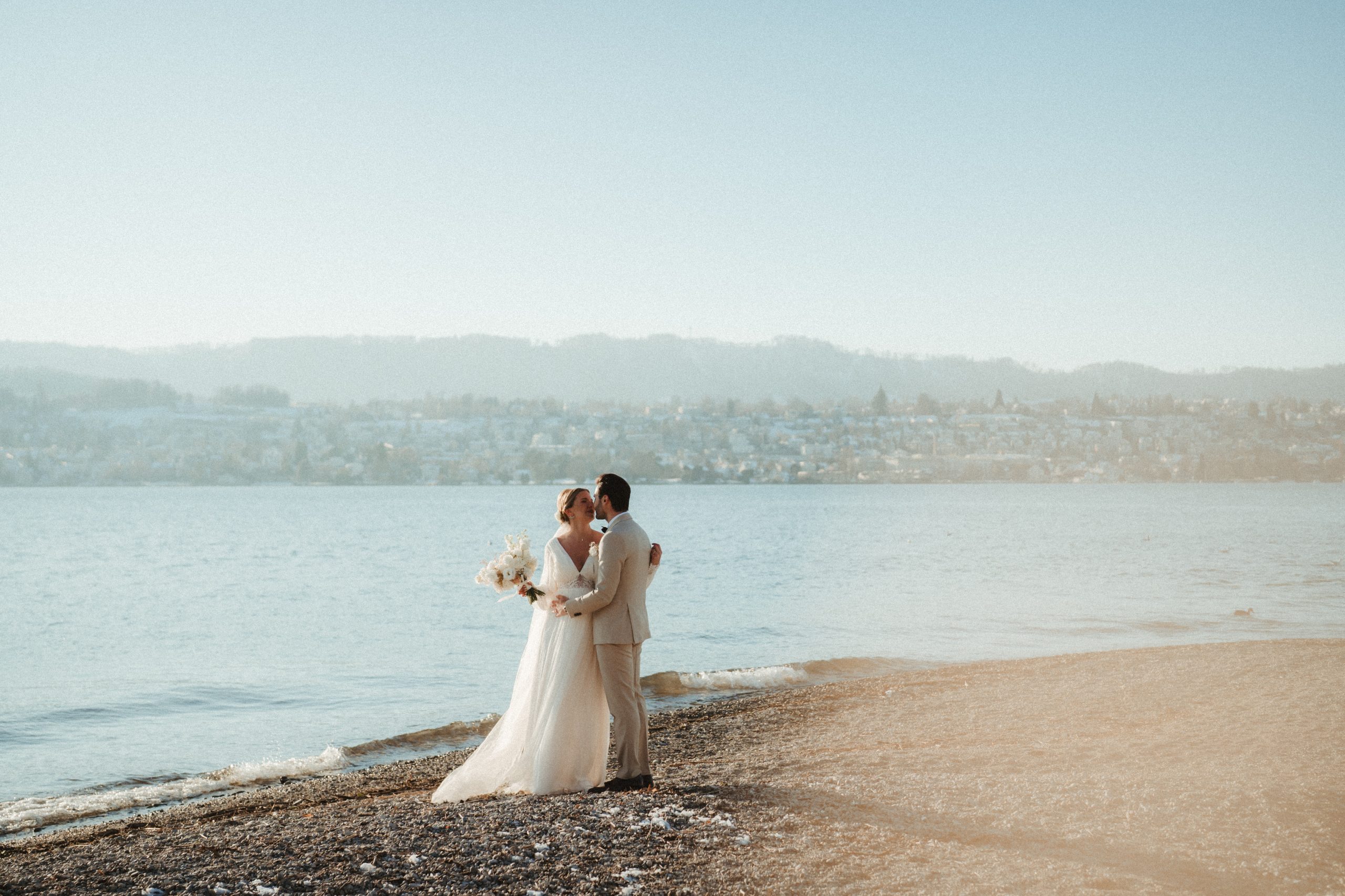 Eine Braut und ein Bräutigam in Hochzeitskleidung umarmen sich an einem friedlichen Seeufer mit einer malerischen Stadt und Hügeln auf der anderen Seite des Wassers unter einem klaren blauen Himmel.