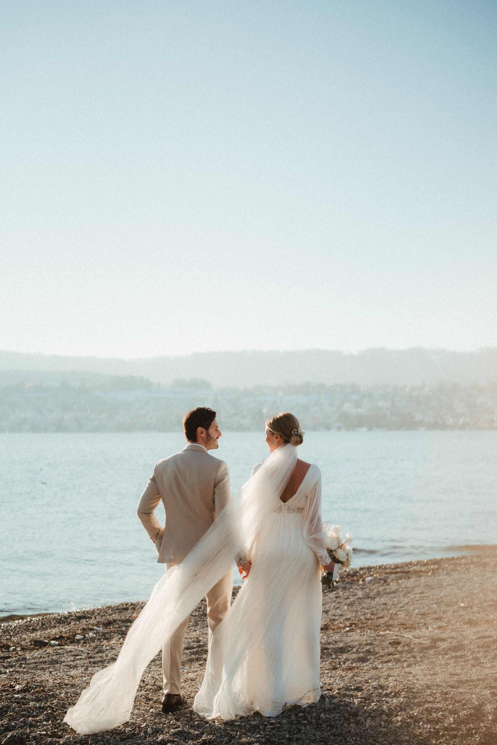 Eine Braut und ein Bräutigam gehen händchenhaltend an einem Kiesstrand entlang. Der lange Schleier der Braut weht in der Brise, und sie schauen sich lächelnd an. Das ruhige Wasser und die fernen Hügel sind unter einem klaren Himmel zu sehen.