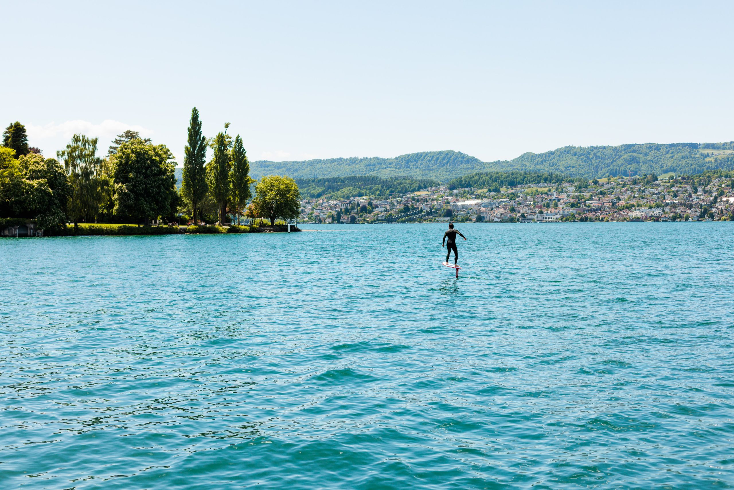 Eine Person in einem Neoprenanzug fährt auf einem Tragflächen-Surfbrett über einen grossen, blauen See mit kräuselndem Wasser, unweit des Boutiquehotels Sonne in Küsnacht, umgeben von grünen Bäumen und fernen Hügeln unter einem klaren Himmel.