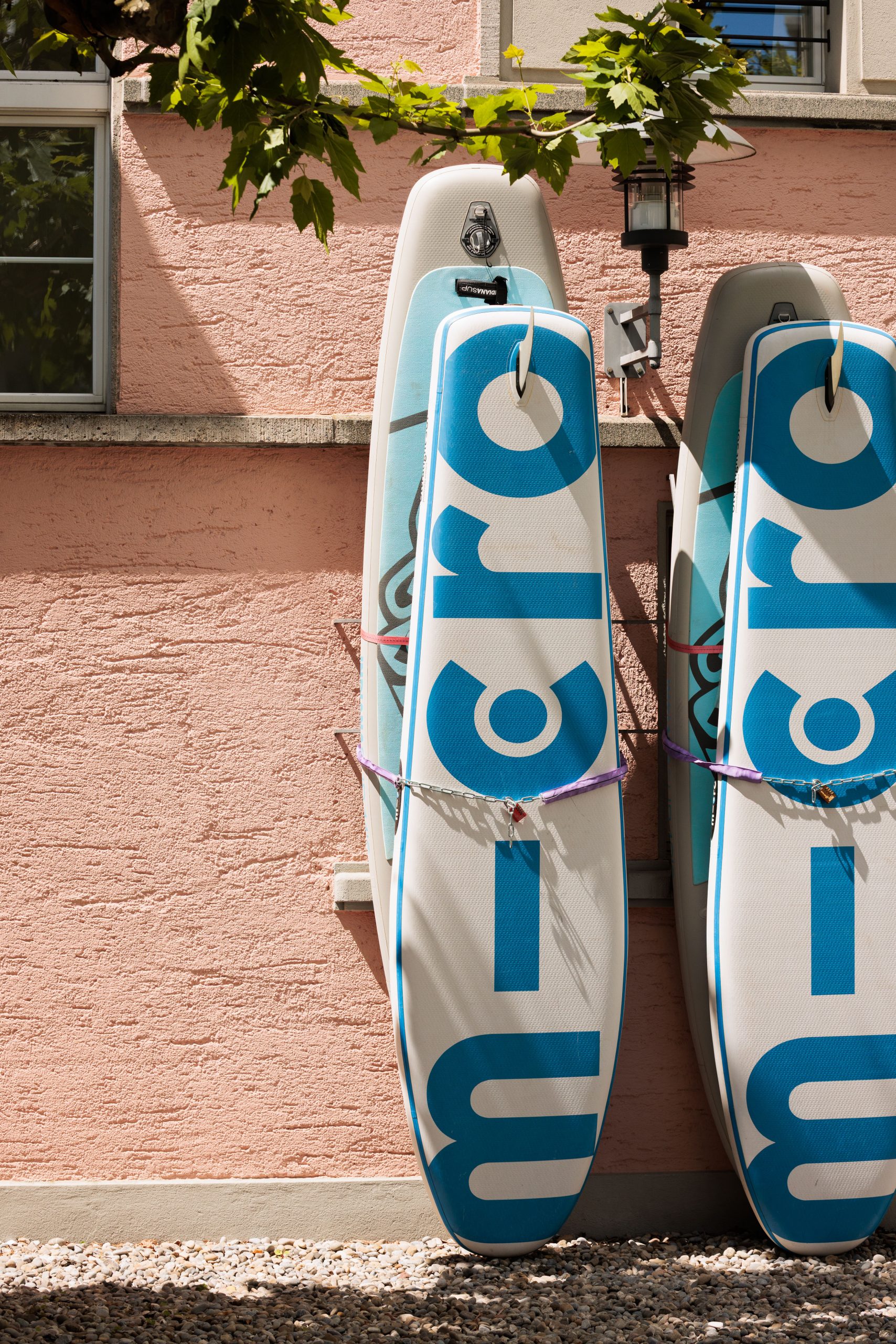 Vor dem Boutiquehotel Hotel am Zürichsee in Küsnacht stehen zwei blau-weiss gestaltete Stand-up-Paddleboards mit dem Schriftzug "micro" aufrecht vor einer sonnenbeschienenen rosa Stuckwand, darüber sind belaubte Äste zu sehen.