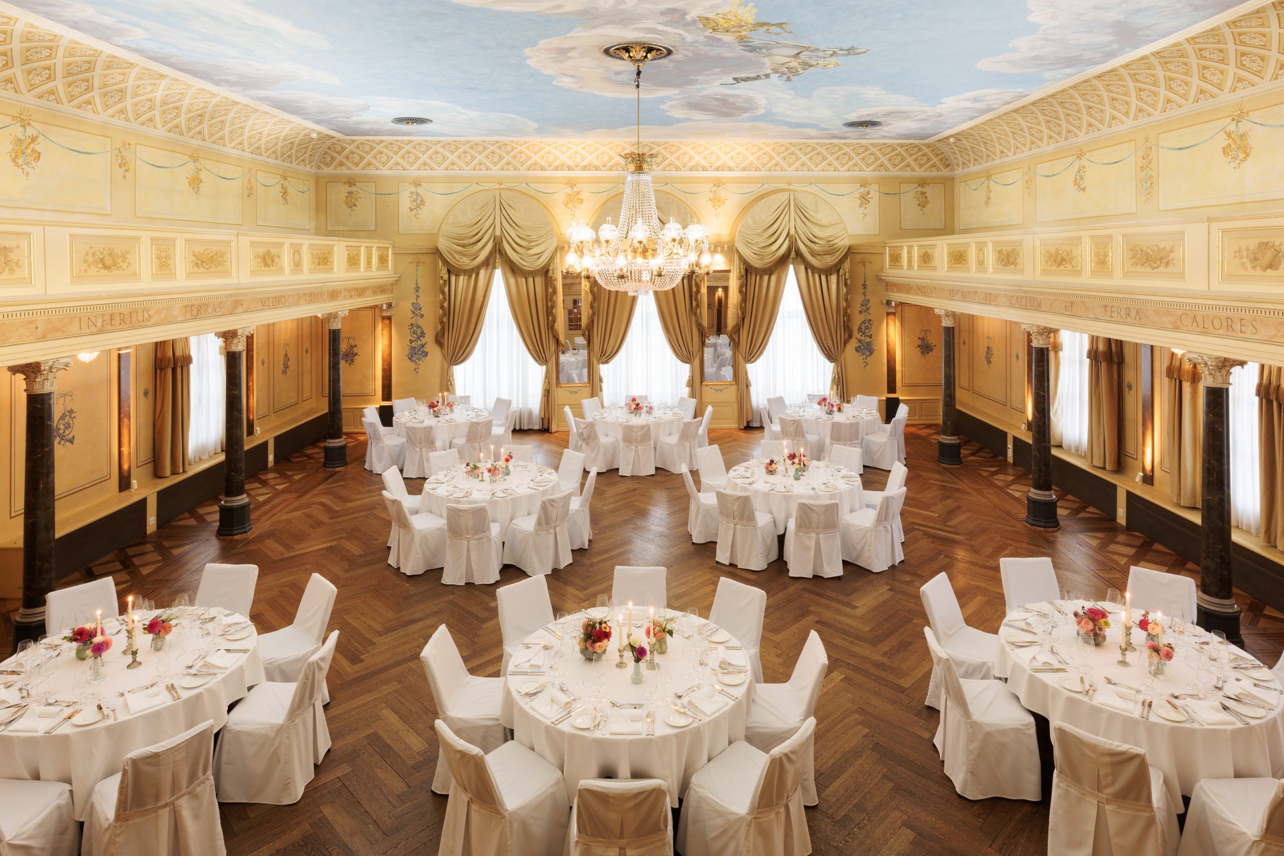 Eleganter Festsaal und Eventlocation am Zürichsee mit verschnörkelten Wänden, einer bemalten Decke und hohen, mit Goldvorhängen drapierten Fenstern. Runde Tische mit weißen Tüchern und Blumenkränzen sind für eine formelle Veranstaltung gedeckt, umgeben von weiß gedeckten Stühlen.