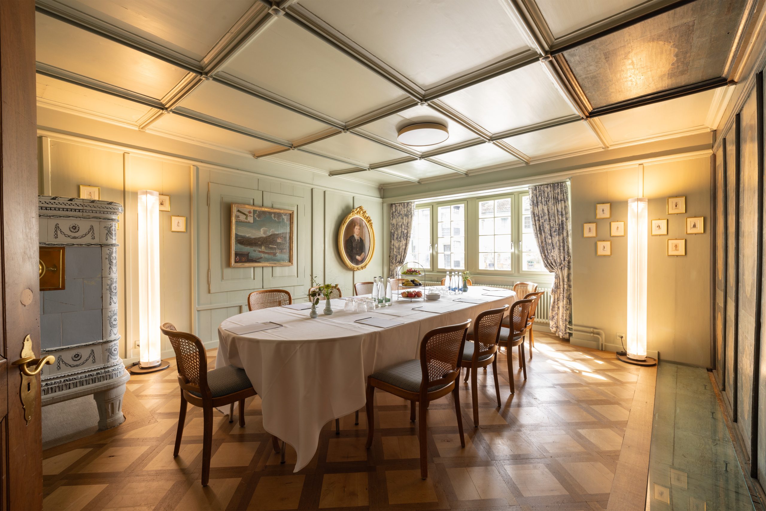 Ein lichtdurchfluteter, eleganter Speisesaal mit einem langen Tisch, der für ein Treffen oder eine Mahlzeit gedeckt ist, umgeben von Holzstühlen, klassischen Porträts an den Wänden, hohen Fenstern mit Vorhängen und warmer Beleuchtung - perfekt für ein unvergessliches Eventraeume.