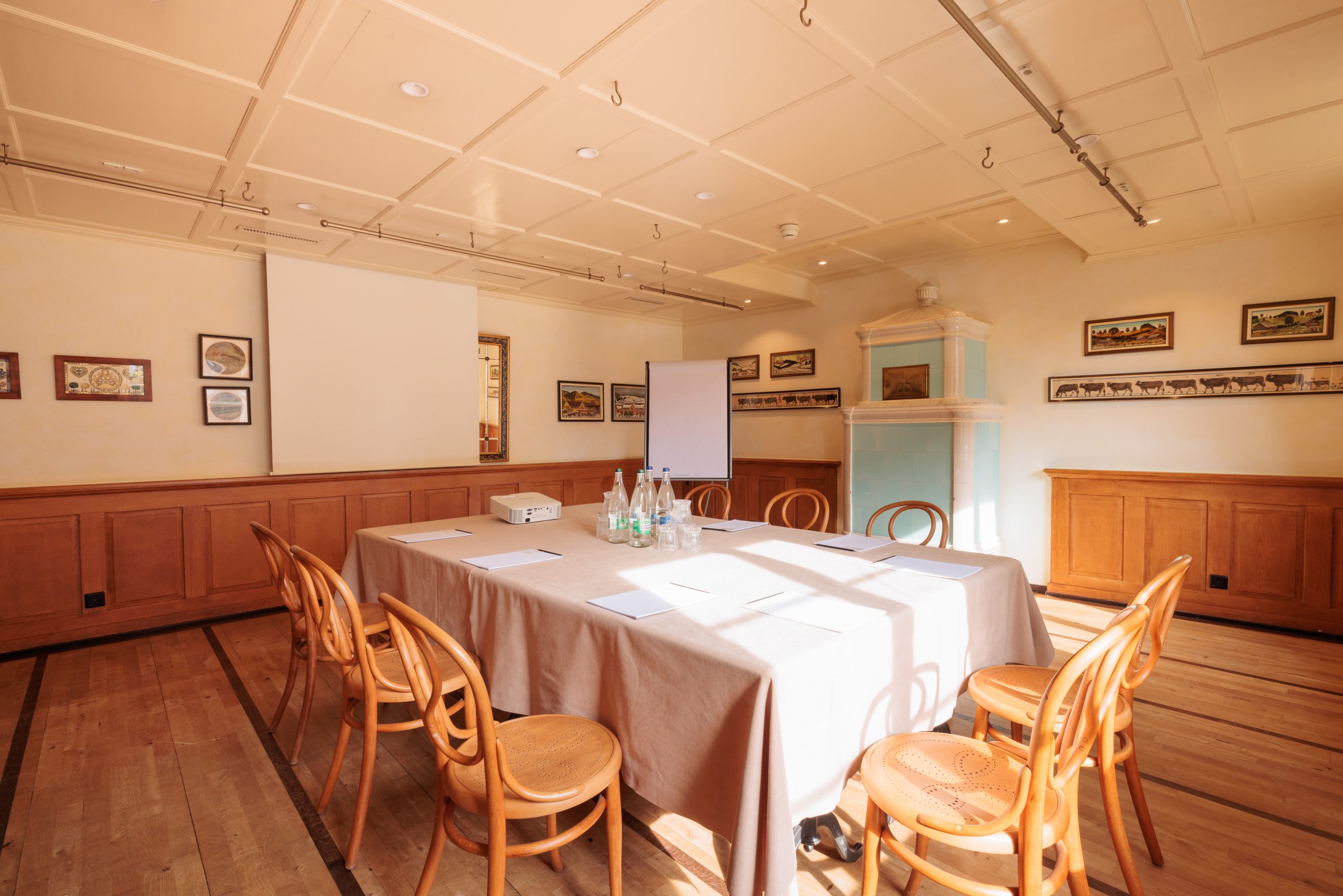 Ein sonnendurchfluteter Besprechungsraum mit einem rechteckigen Tisch, umgeben von Holzstühlen, Papieren und Flaschen auf dem Tisch, einem Projektor, einer Leinwand und gerahmten Presse-Kunstwerken an den Wänden.