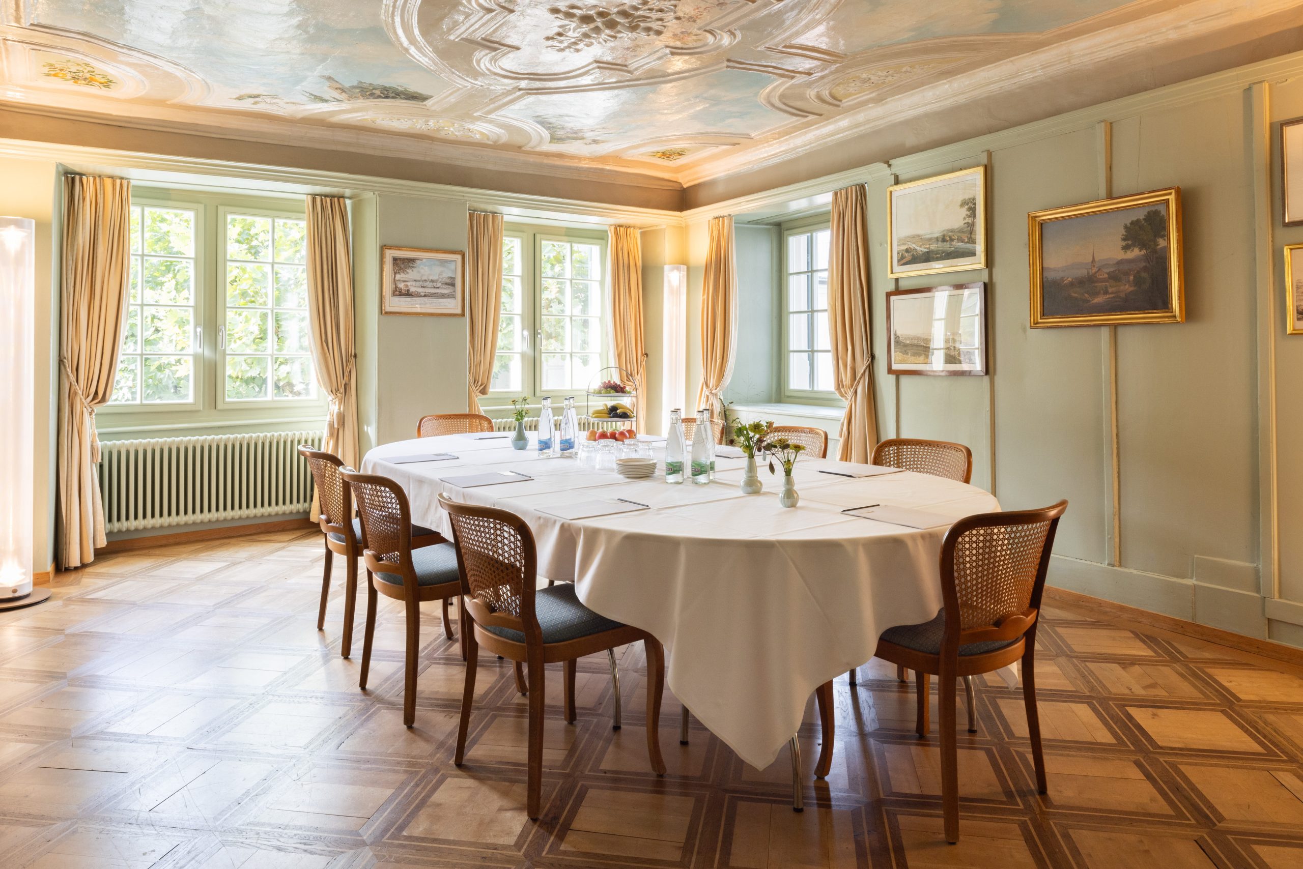 Elegantes Esszimmer mit einem quadratischen, mit einer weißen Tischdecke bedeckten Tisch, der für eine Besprechung mit Glasflaschen, Gläsern, Papieren und Stiften gedeckt ist. Der Raum hat große Fenster, helle Vorhänge, Kunstwerke und verschnörkelte Deckendetails.