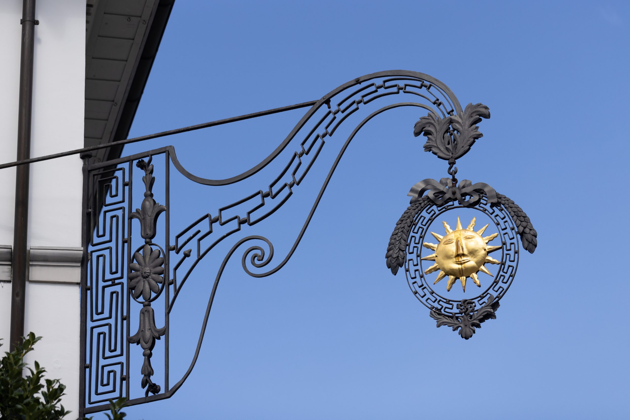 Ein dekoratives schmiedeeisernes Schild mit einem goldenen Sonnengesicht hängt an einem Gebäude vor einem klaren blauen Himmel. Das Schild weist verschnörkelte Muster und blattartige Details auf.