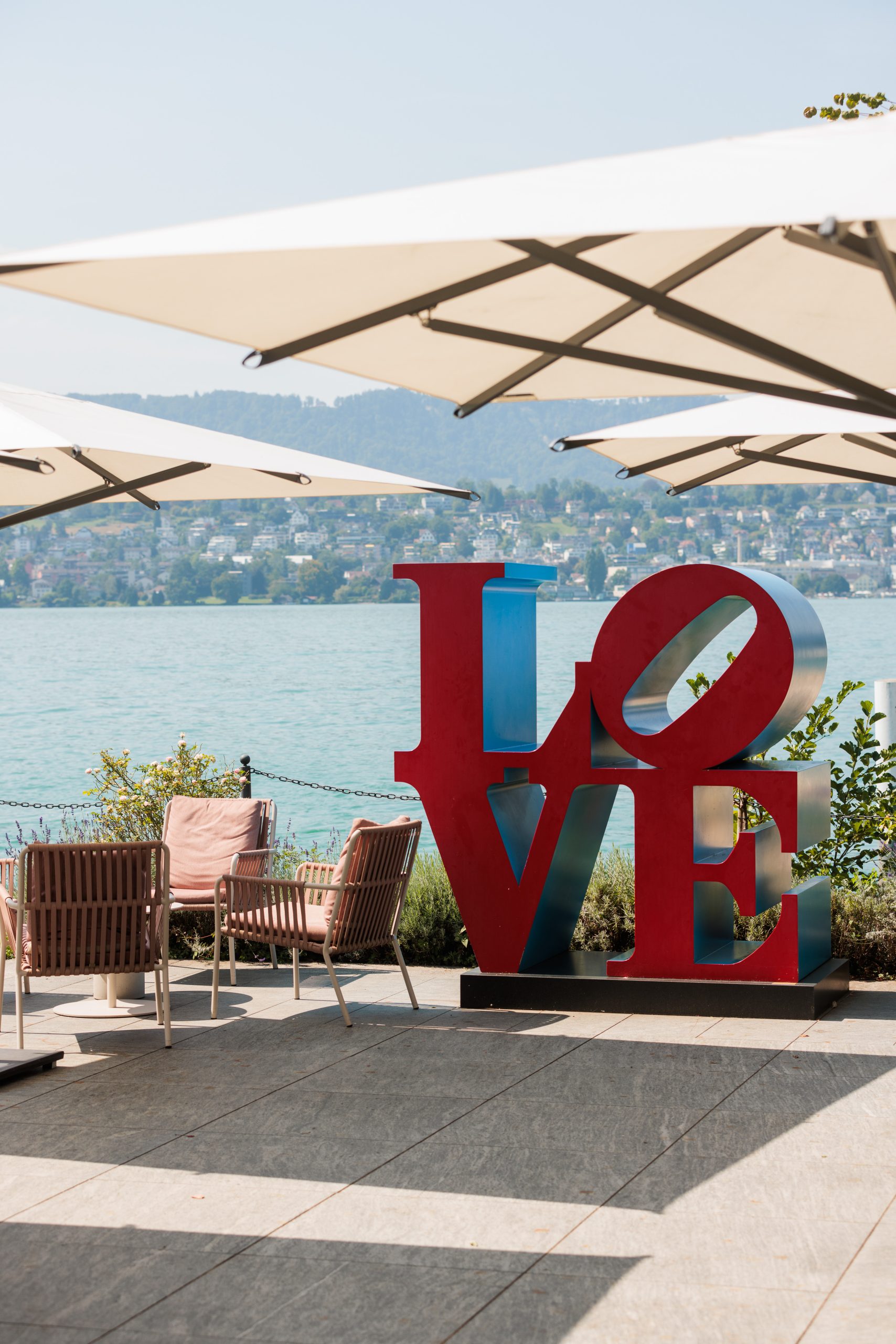 Eine große rote und blaue LOVE-Skulptur steht auf einer Terrasse an einem See, mit Stühlen, Tischen und Sonnenschirmen der Presse im Freien über dem Wasser. Im Hintergrund sind über das Wasser hinweg Hügel und Gebäude zu sehen.