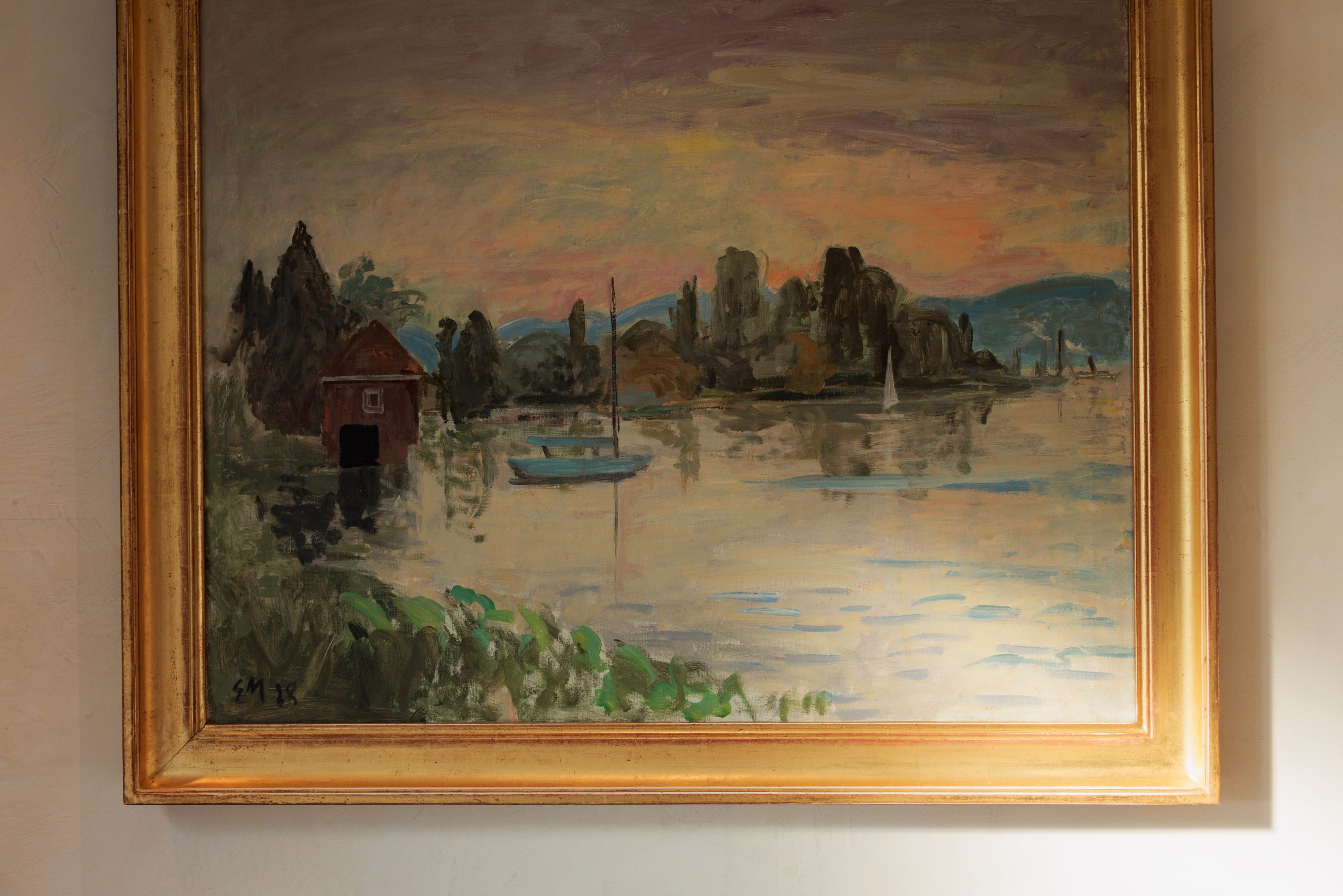 Ein gerahmtes Gemälde mit einer Seeszene bei Sonnenuntergang, inspiriert von der Kunst und Geschichte rund um das Boutiquehotel Sonne, zeigt ein rotes Bootshaus, Segelboote auf dem Wasser und Bäume mit fernen Hügeln unter einem farbenfrohen Himmel.