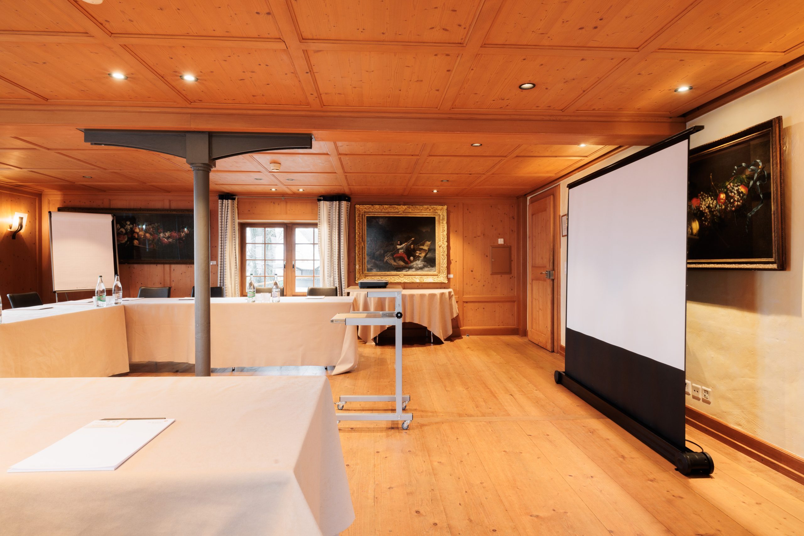 Ein Konferenzraum mit hölzernen Wänden und Böden, ausgestattet mit Tischen und Stühlen, einem Projektor, Flipchart, Wasserflaschen und Bildern an den Wänden - ideal für Presse-Veranstaltungen. Durch ein Fenster mit eleganten Vorhängen fällt natürliches Licht ein.