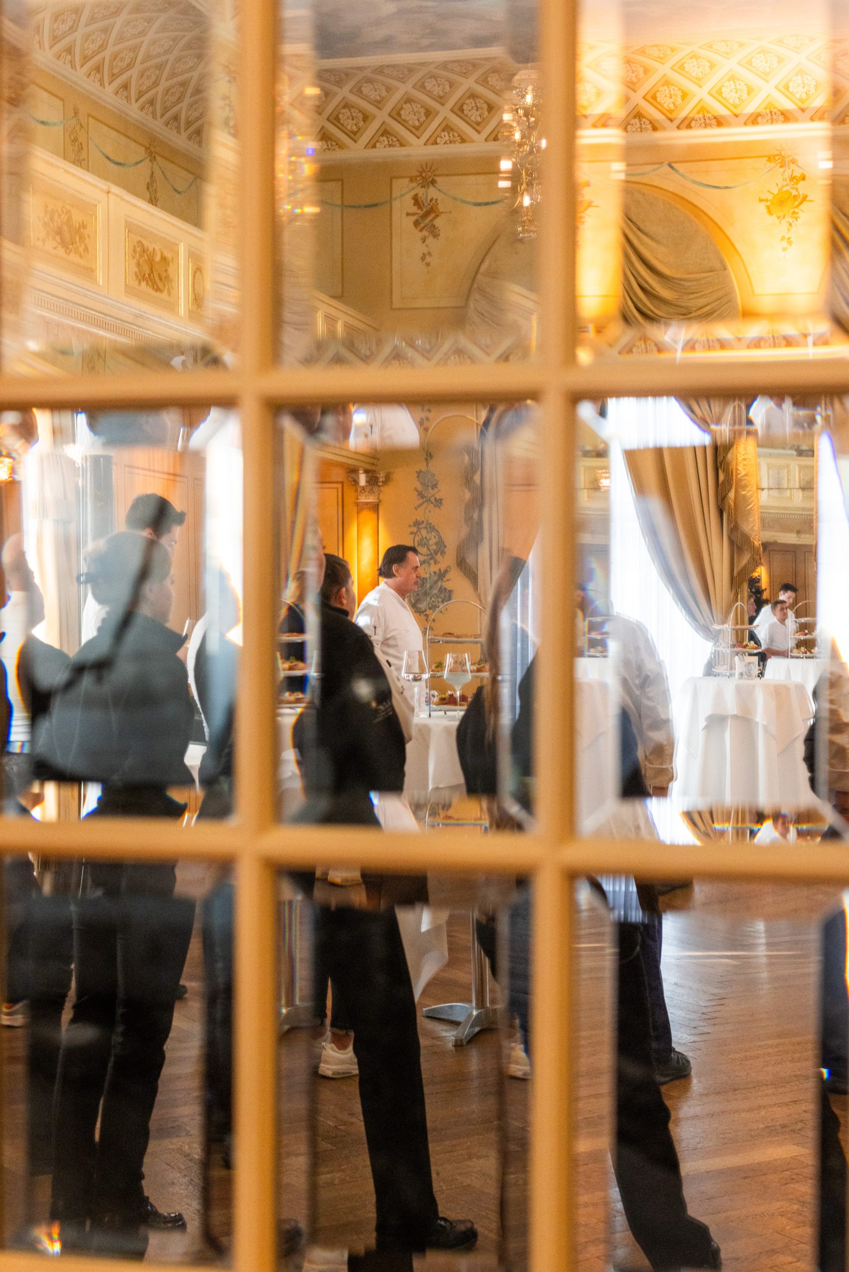 Eine Gruppe von Menschen steht und unterhält sich in einem eleganten, verzierten Raum mit goldenen Wänden und gewölbten Fenstern, die durch eine Glastür zu sehen sind und sowohl die Pracht im Inneren als auch den exklusiven Reiz der Spitzenjobs der Hotellerie widerspiegeln.