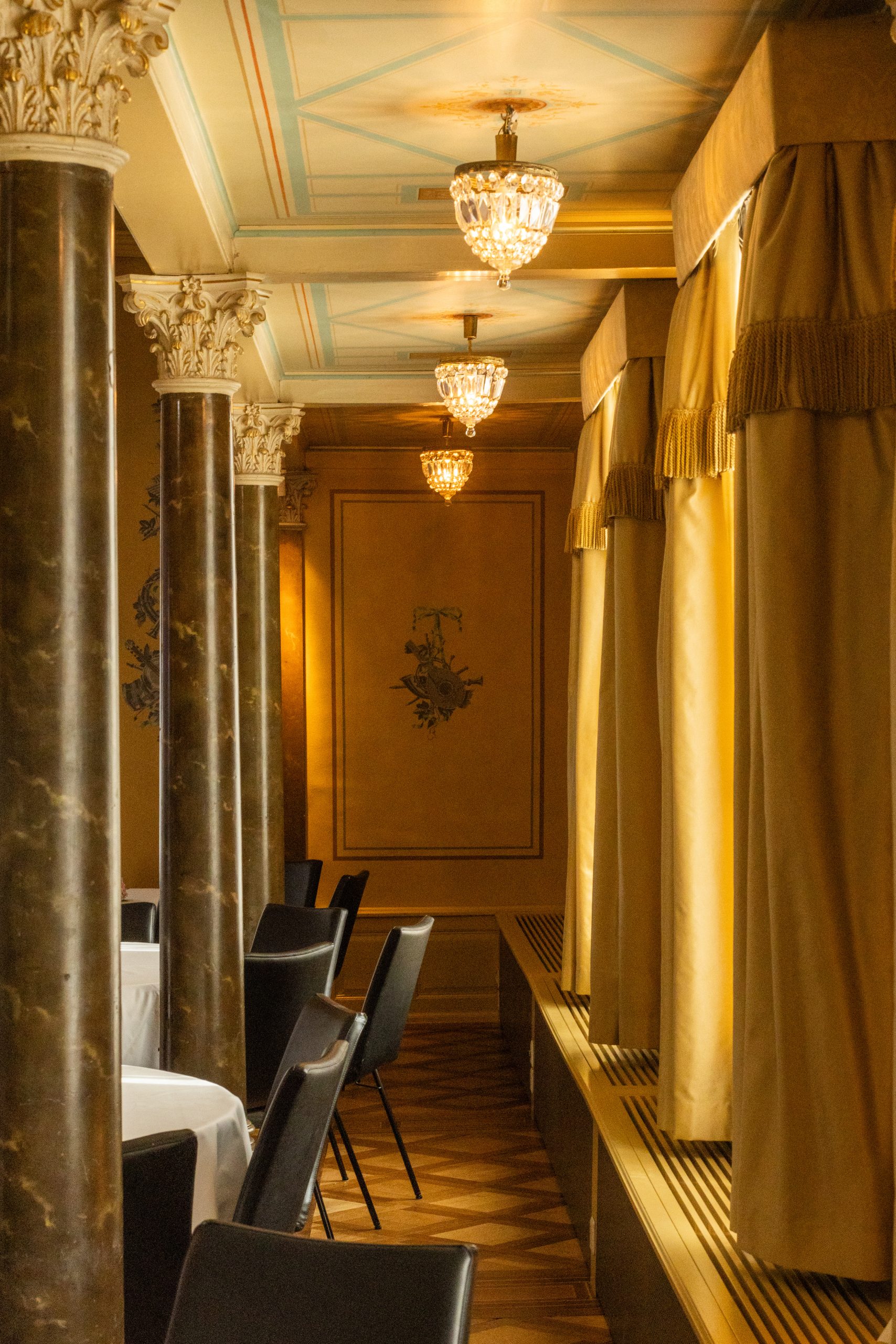 Elegantes Restaurantinterieur mit verschnörkelten Säulen, drapierten Fenstern, Kristalllüstern und weiß gedeckten Tischen im Presse-Stil; warme Beleuchtung schafft eine gemütliche und anspruchsvolle Atmosphäre.