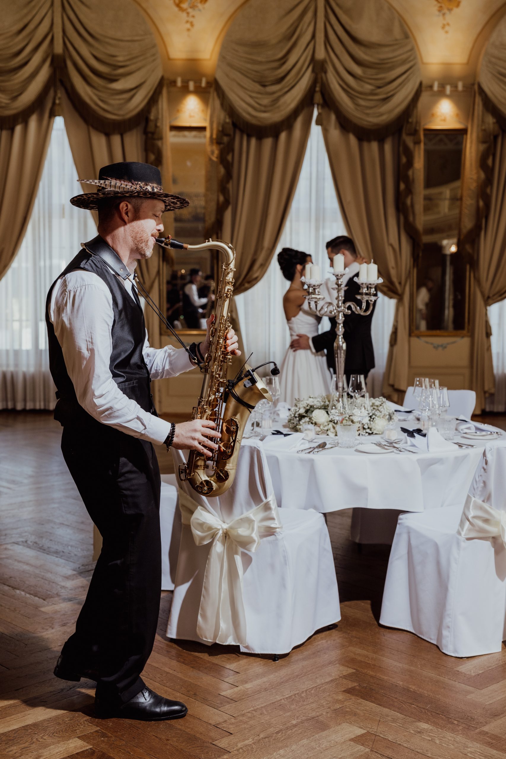 Ein Musiker mit Hut spielt Saxophon neben einem geschmückten Tisch, während sich Braut und Bräutigam im Hintergrund eines eleganten, warm beleuchteten Raums mit großen Fenstern und drapierten Vorhängen umarmen.