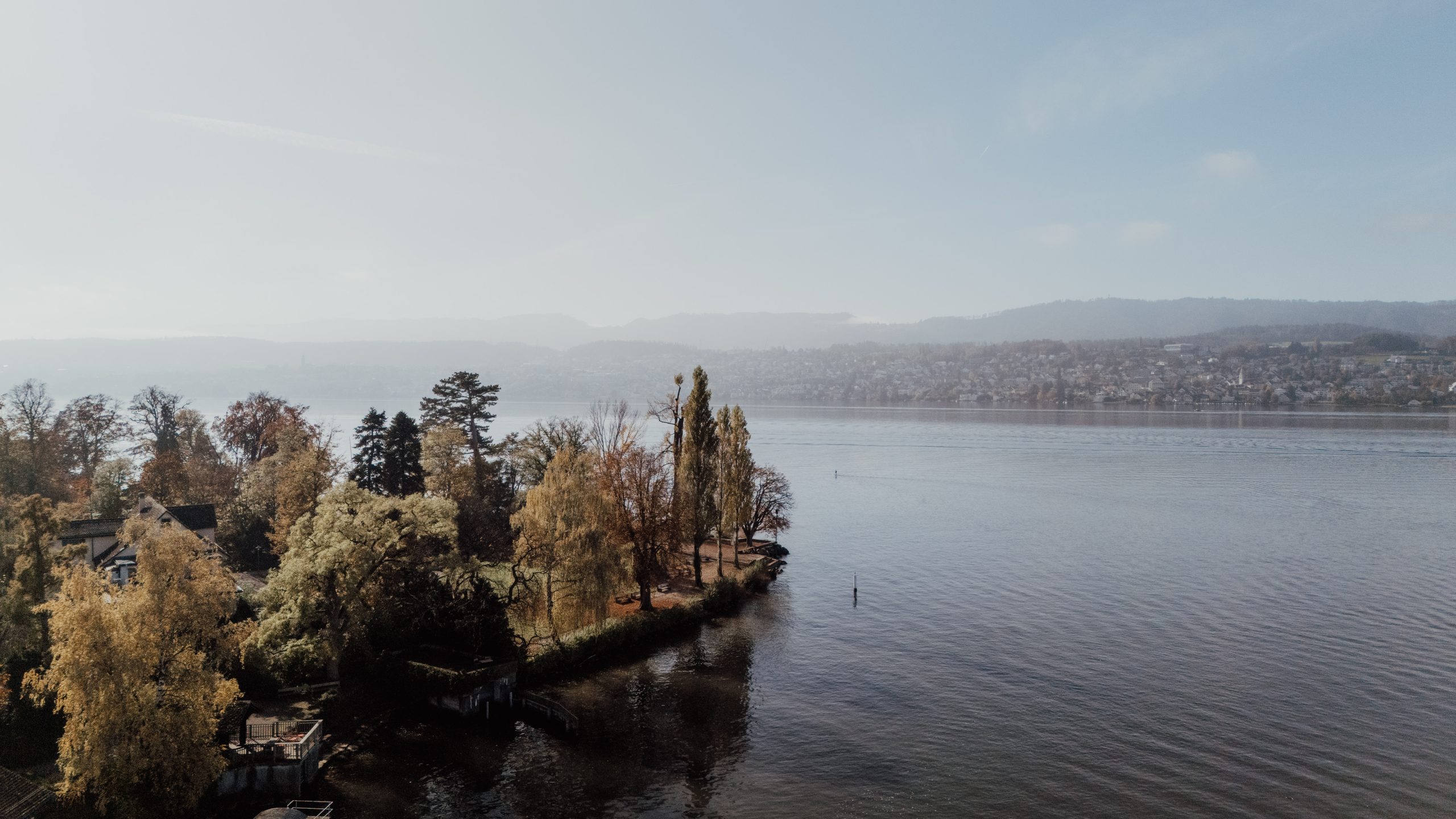 Ein ruhiger See mit sanftem Plätschern, gesäumt von herbstlich gefärbten Bäumen auf einer kleinen Halbinsel. In der Ferne sind Häuser und Hügel unter einem dunstigen, blassblauen Himmel zu sehen.