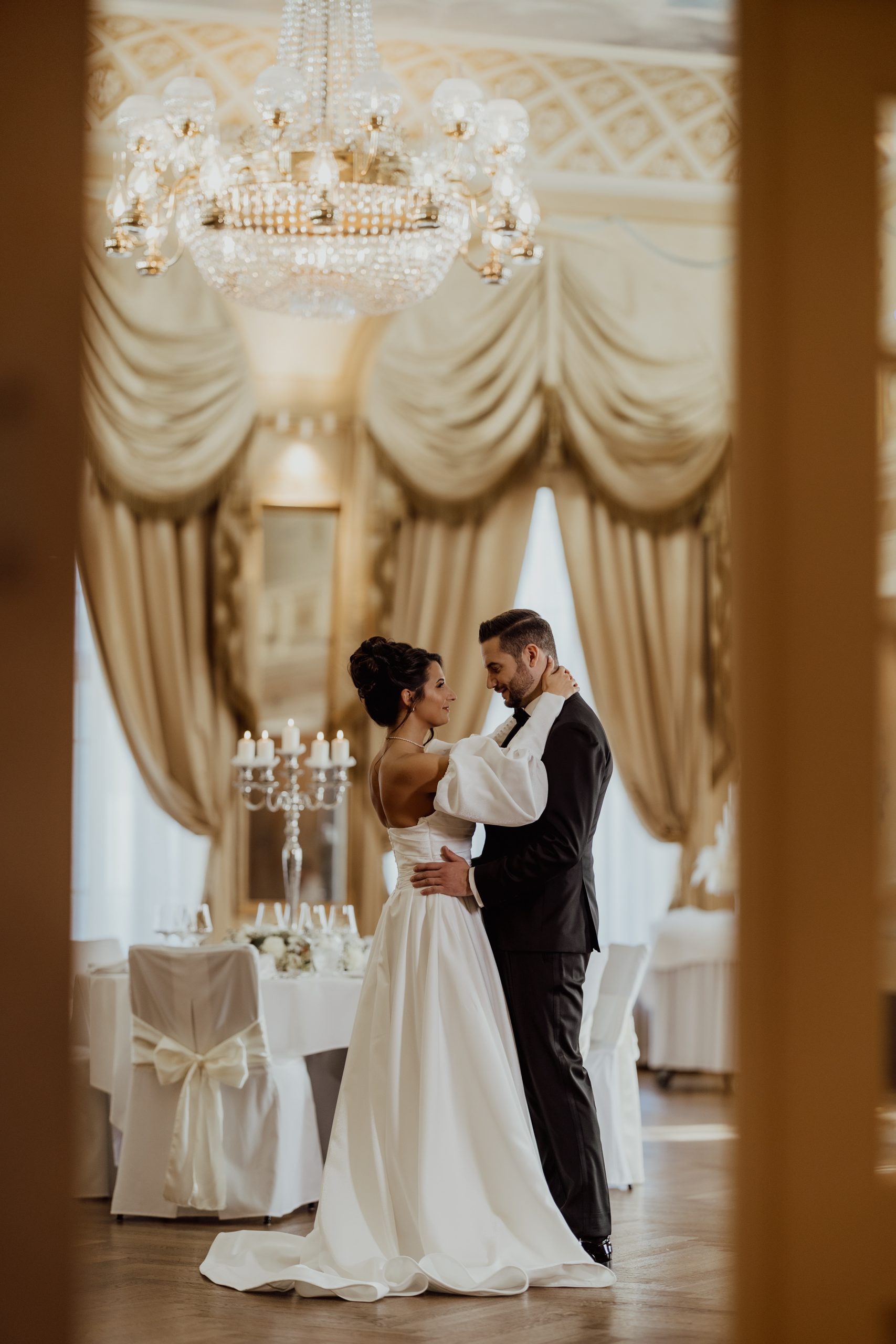 Eine Braut und ein Bräutigam tanzen einen romantischen Tanz in einem eleganten, von einem Kronleuchter beleuchteten Raum mit verzierten Vorhängen und dekorierten Tischen im Hintergrund. Die Braut trägt ein weißes Kleid, und der Bräutigam trägt einen schwarzen Anzug.
