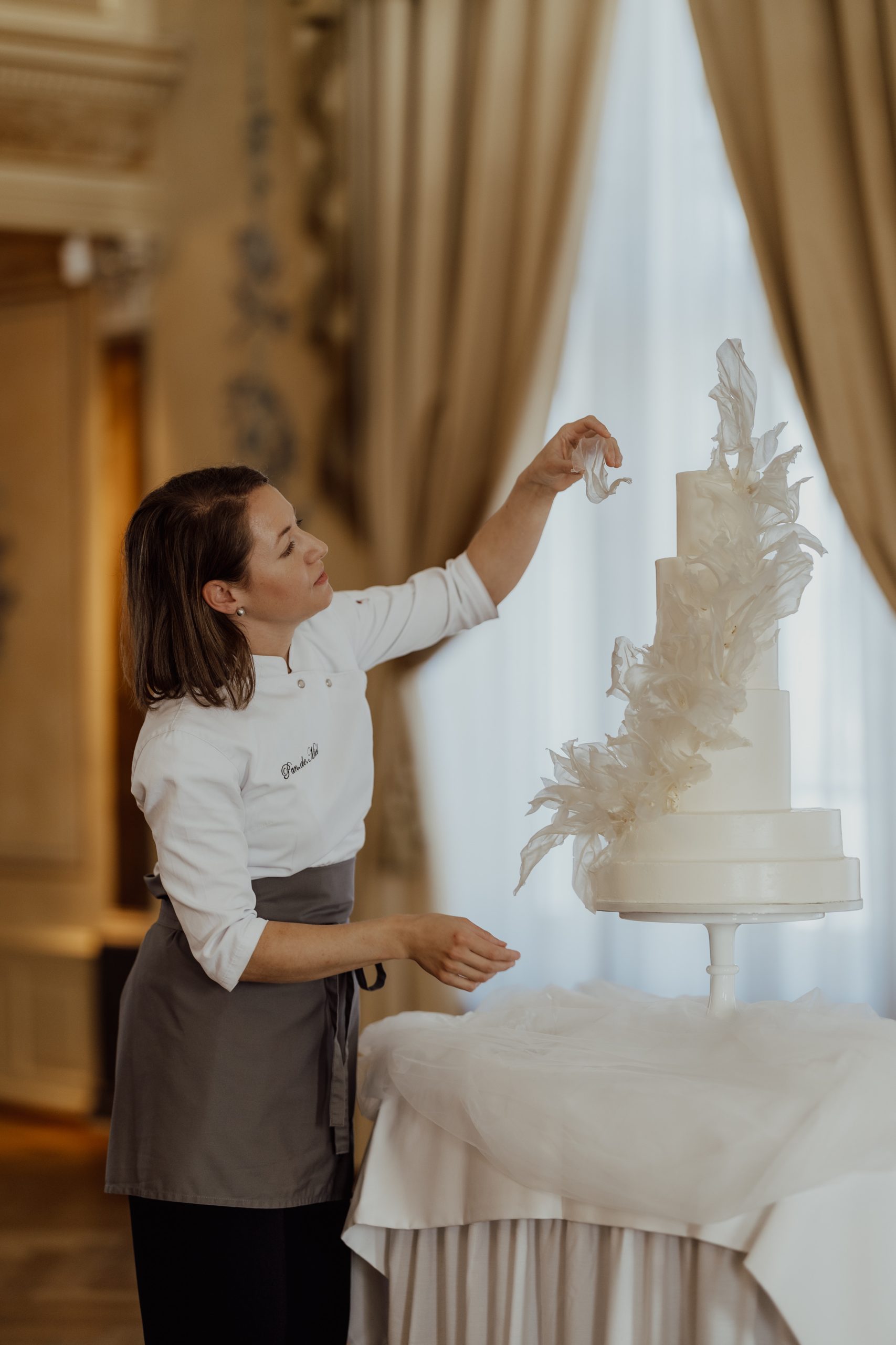 Eine Frau in einer Kochuniform verziert in einem verzierten, sanft beleuchteten Raum eine hohe, elegante weiße Hochzeitstorte mit zarten weißen Zuckerdekorationen.