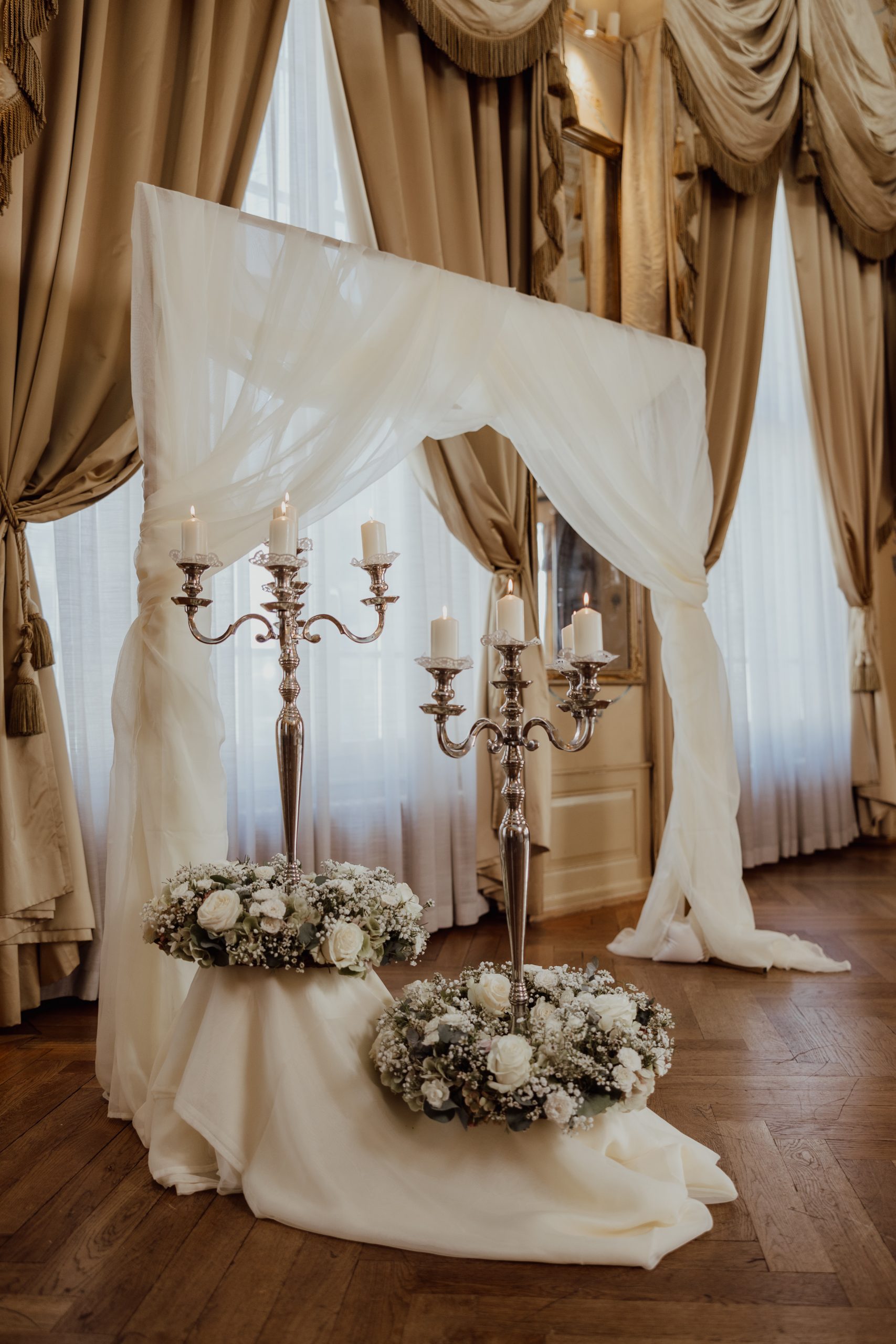 Eleganter Hochzeitsbogen mit fließendem weißem Stoff, silbernen Leuchtern mit brennenden Kerzen und Blumenarrangements mit weißen Blumen und Grünzeug in einem Raum mit hohen Fenstern und goldenen Vorhängen.