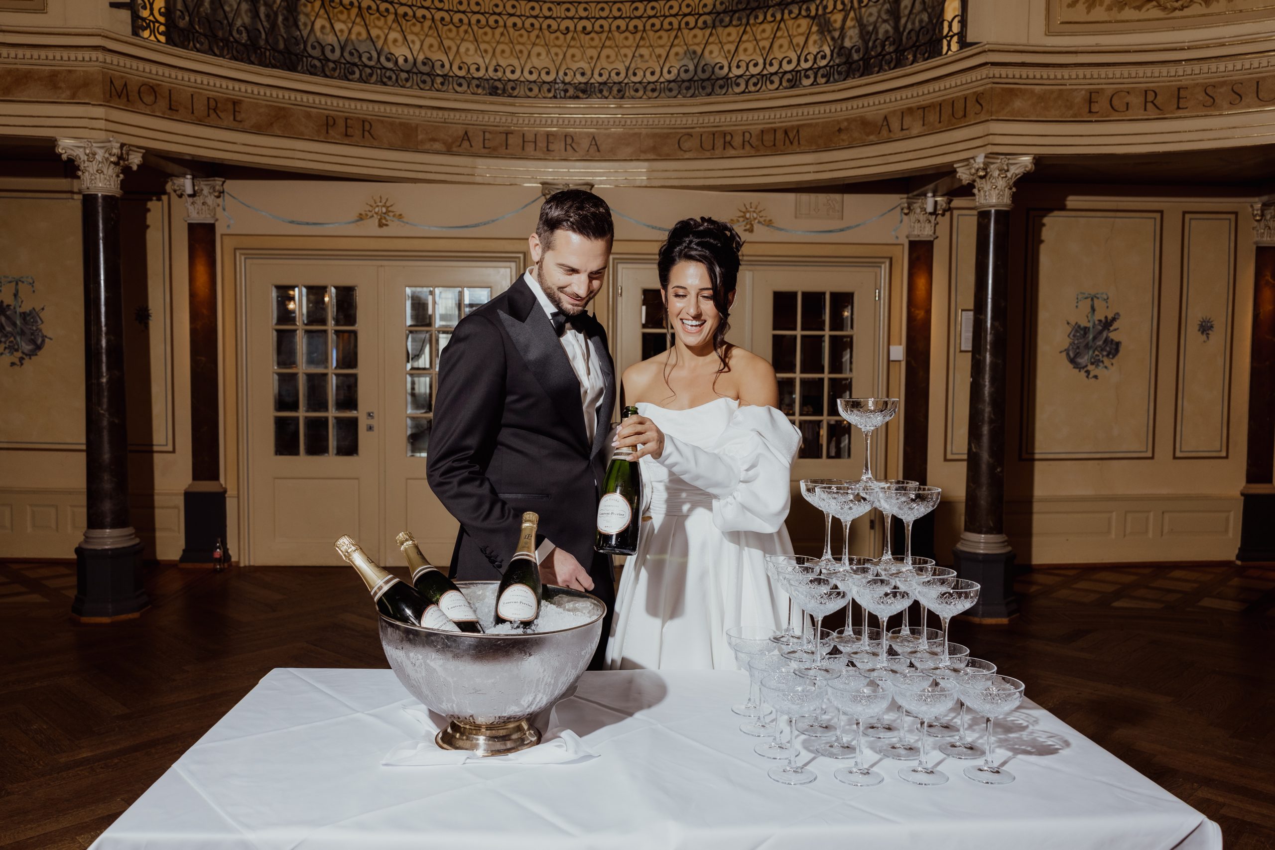 Ein formal gekleidetes Brautpaar lächelt, während der Bräutigam in einem eleganten, verzierten Raum Champagner in einen Turm von Gläsern auf einem weißen Tisch mit Champagnerflaschen in einem Eiskübel gießt.