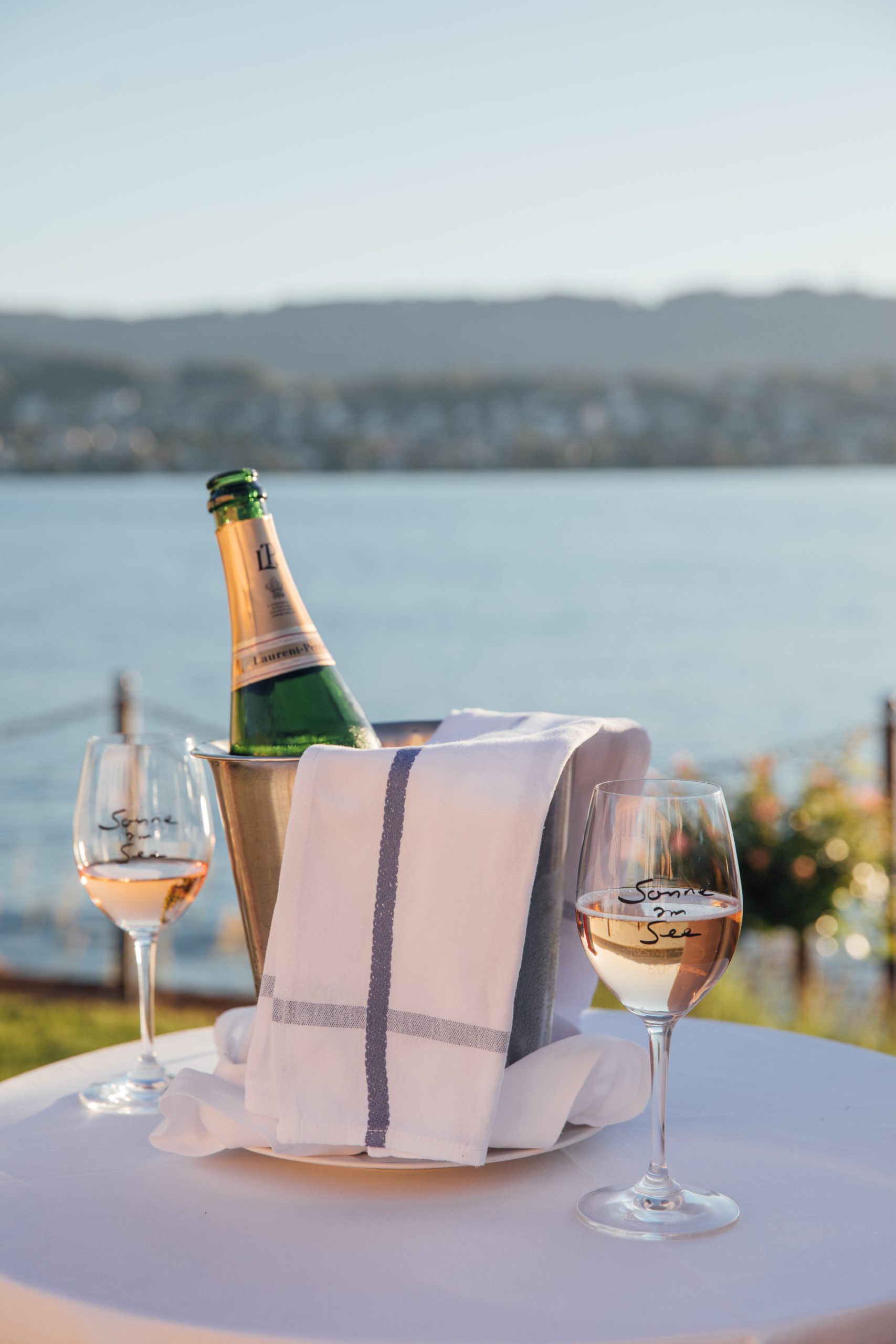 Eine Flasche Champagner in einem Eiskübel, über den ein weißes Handtuch drapiert ist, und zwei Presse-Gläser mit Rosé auf einem Tisch mit Blick auf einen See und die Berge im Hintergrund.