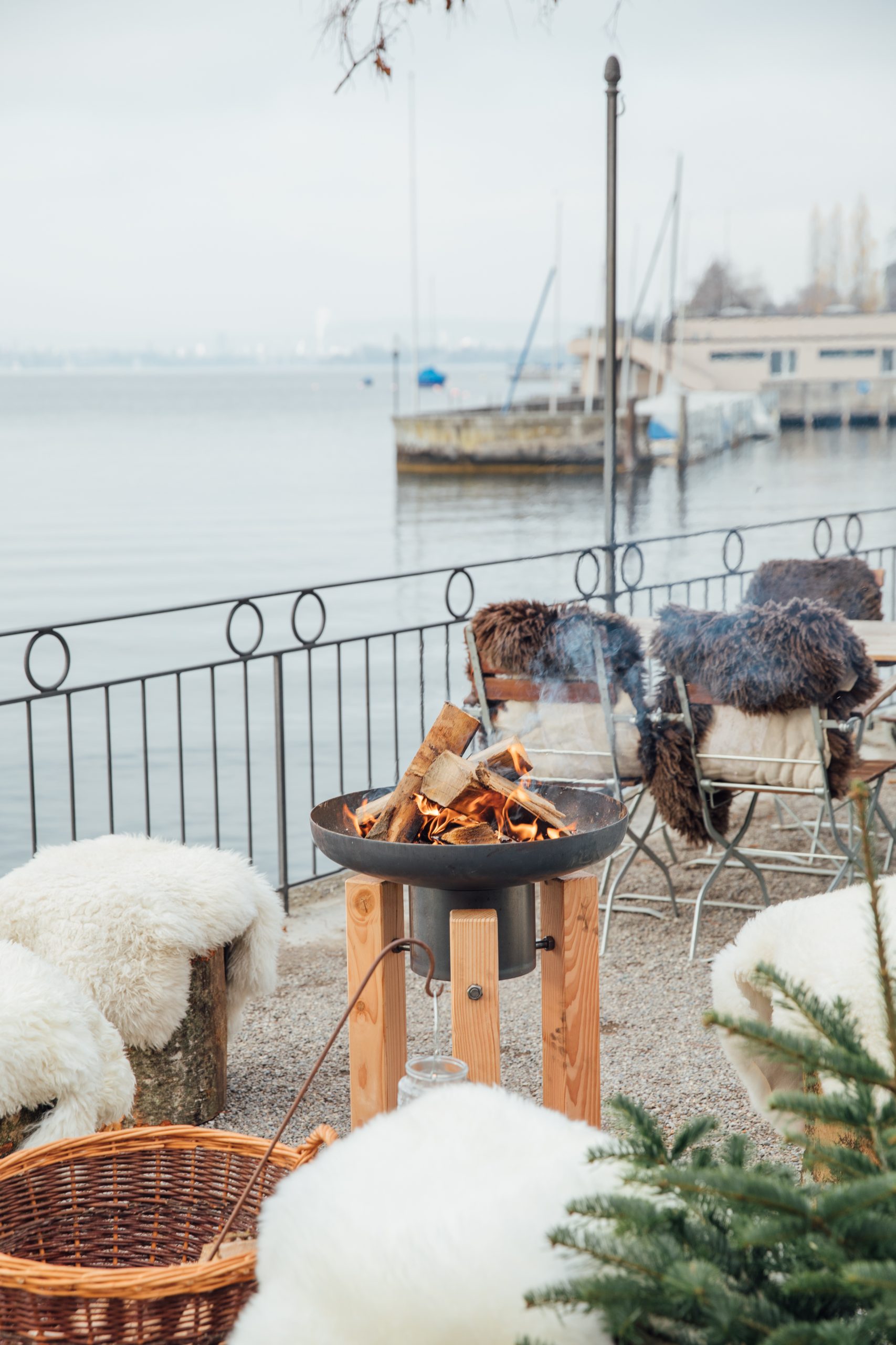 Im Restaurant Seegarten gibt es einen gemütlichen Sitzplatz im Freien am Wasser mit einer Feuerstelle mit brennendem Holz, fellbezogenen Stühlen und einem Weidenkorb, während im Hintergrund Boote und ein Pier zu sehen sind.