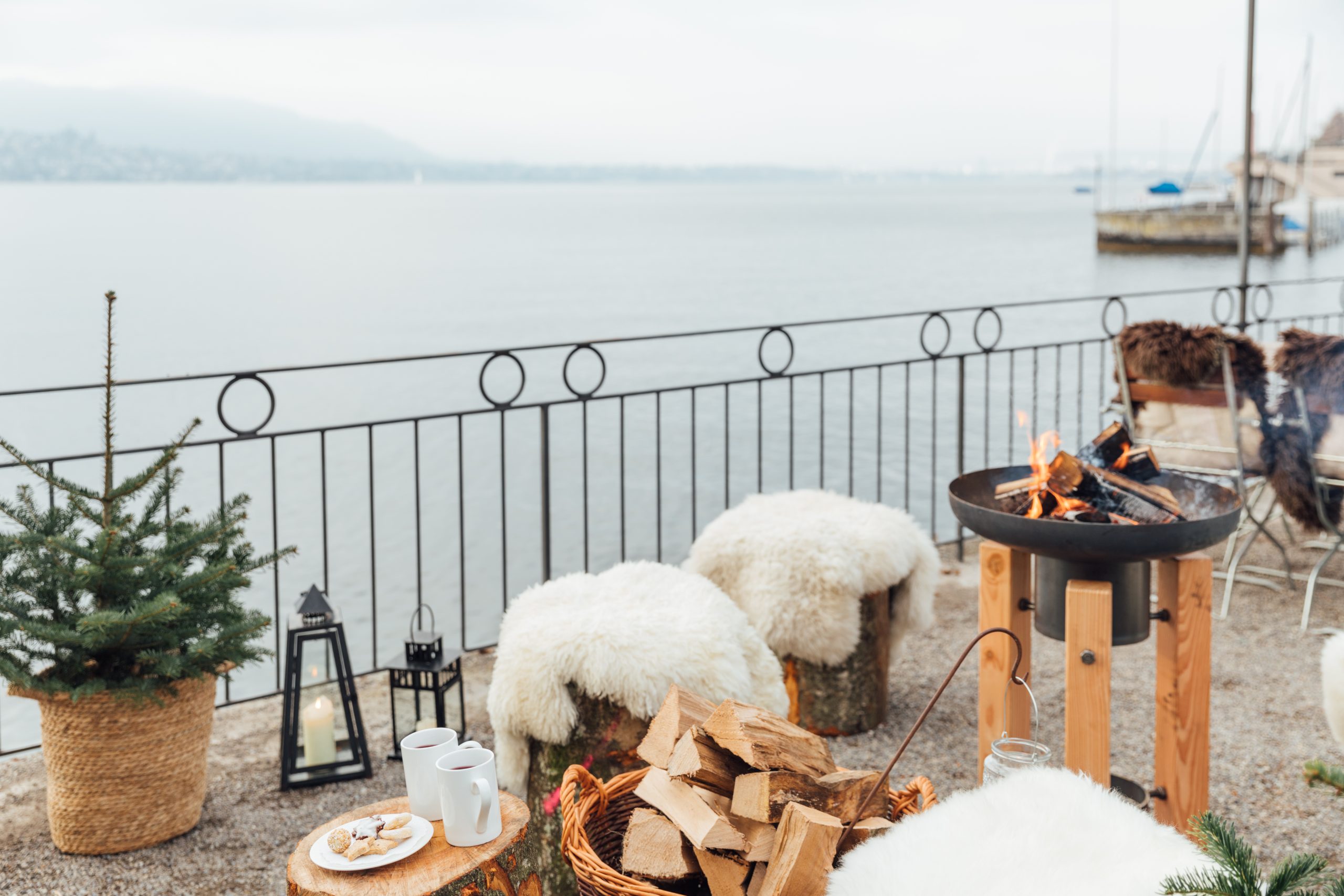 Le restaurant Küsnacht, situé au bord du lac, propose des sièges confortables en plein air, avec des tabourets recouverts de fourrure, un foyer, un sapin dans un panier, des mugs, des biscuits sur la table et du bois de chauffage empilé, le tout à l'intérieur d'une clôture en métal.