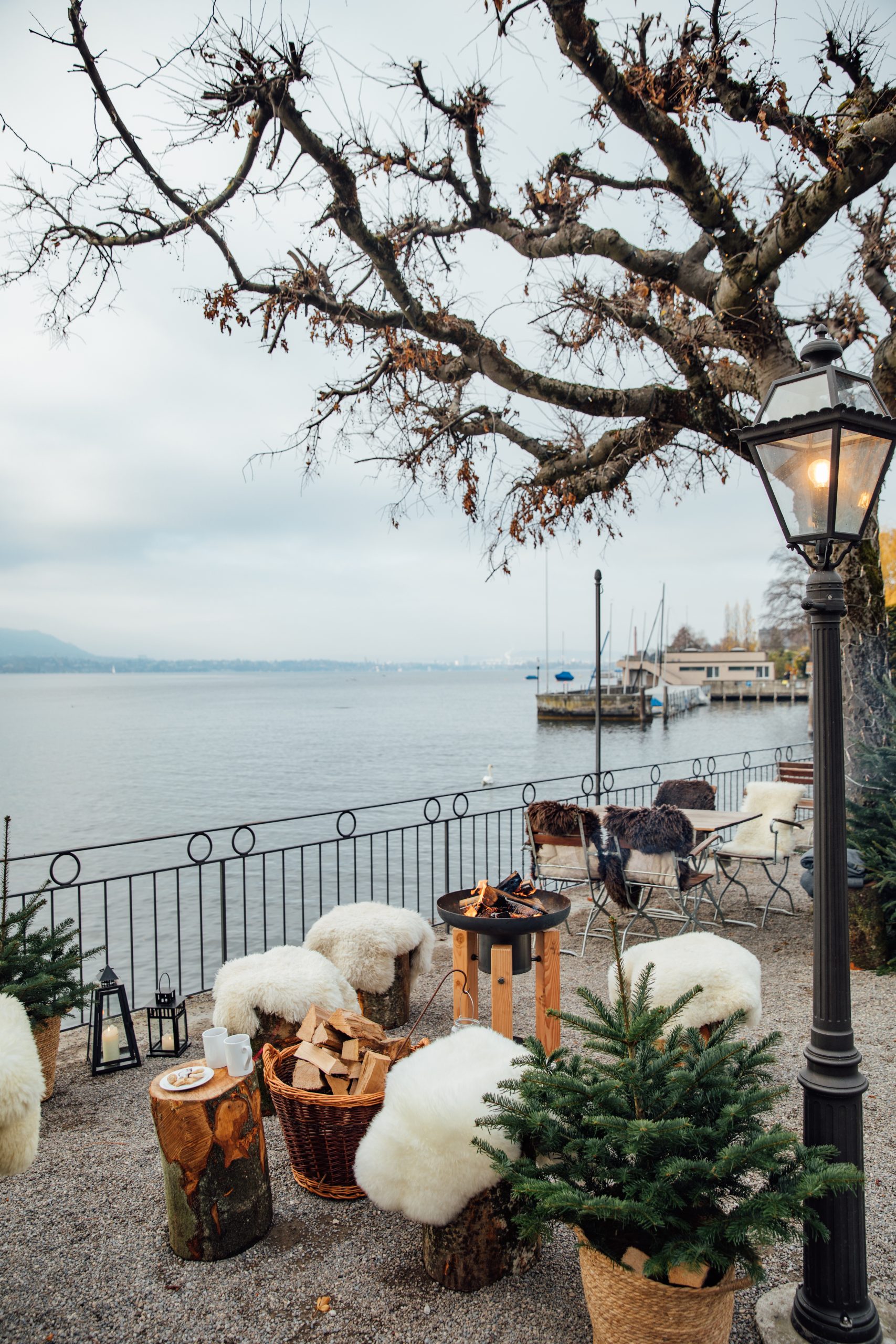 Eine gemütliche Seeterrasse im Restaurant Seegarten mit fellbezogenen Stühlen, einer Feuerstelle, Körben mit Brennholz, kleinen Kiefern und einem blattlosen Baum, mit Blick auf das ruhige Wasser und entfernte Boote an einem bewölkten Tag.