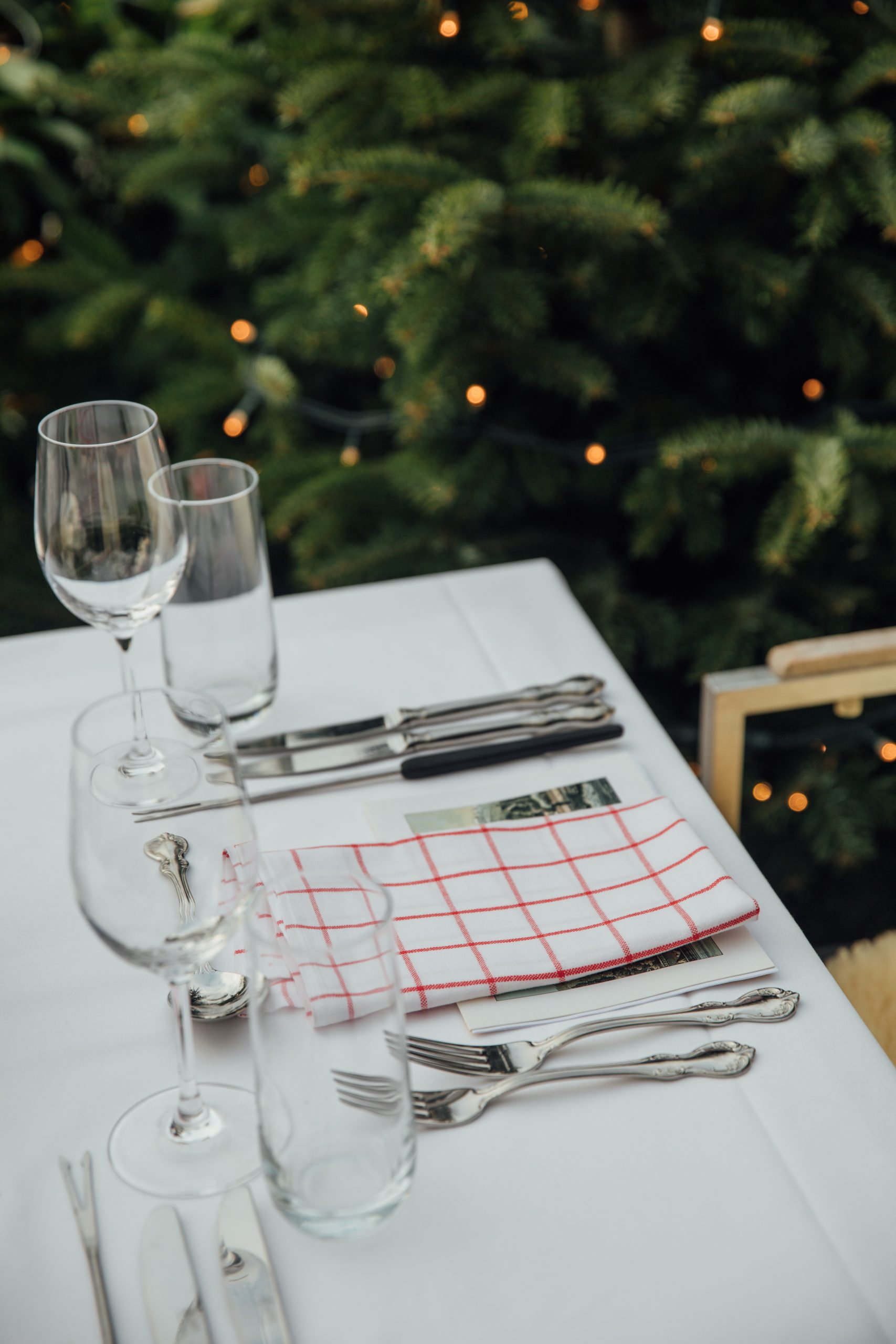 A table set for two with wine glasses, water glasses, silverware, and a red-checkered napkin on a white tablecloth, evokes a cozy evening at a chalet by the lake, with a blurred green Christmas tree with lights in the background.
