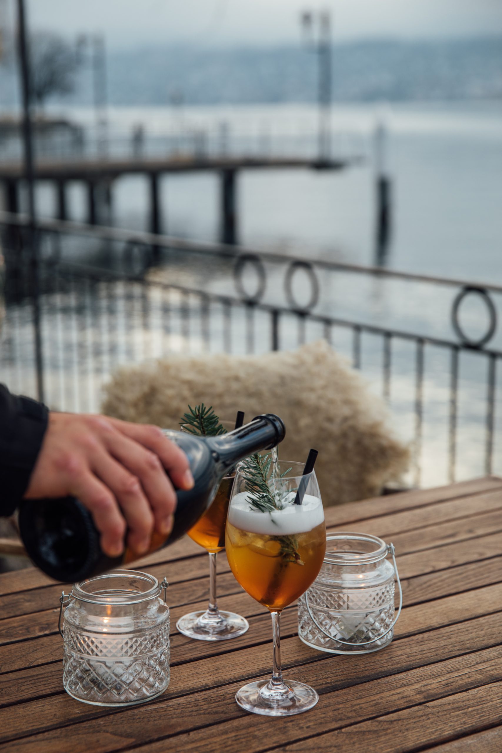 Eine Hand gießt Sekt in zwei Gläser mit Eis und Kräutern im Chalet am See, das auf einem Holztisch am Wasser steht, mit Kerzenschein und einem Pier im Hintergrund.