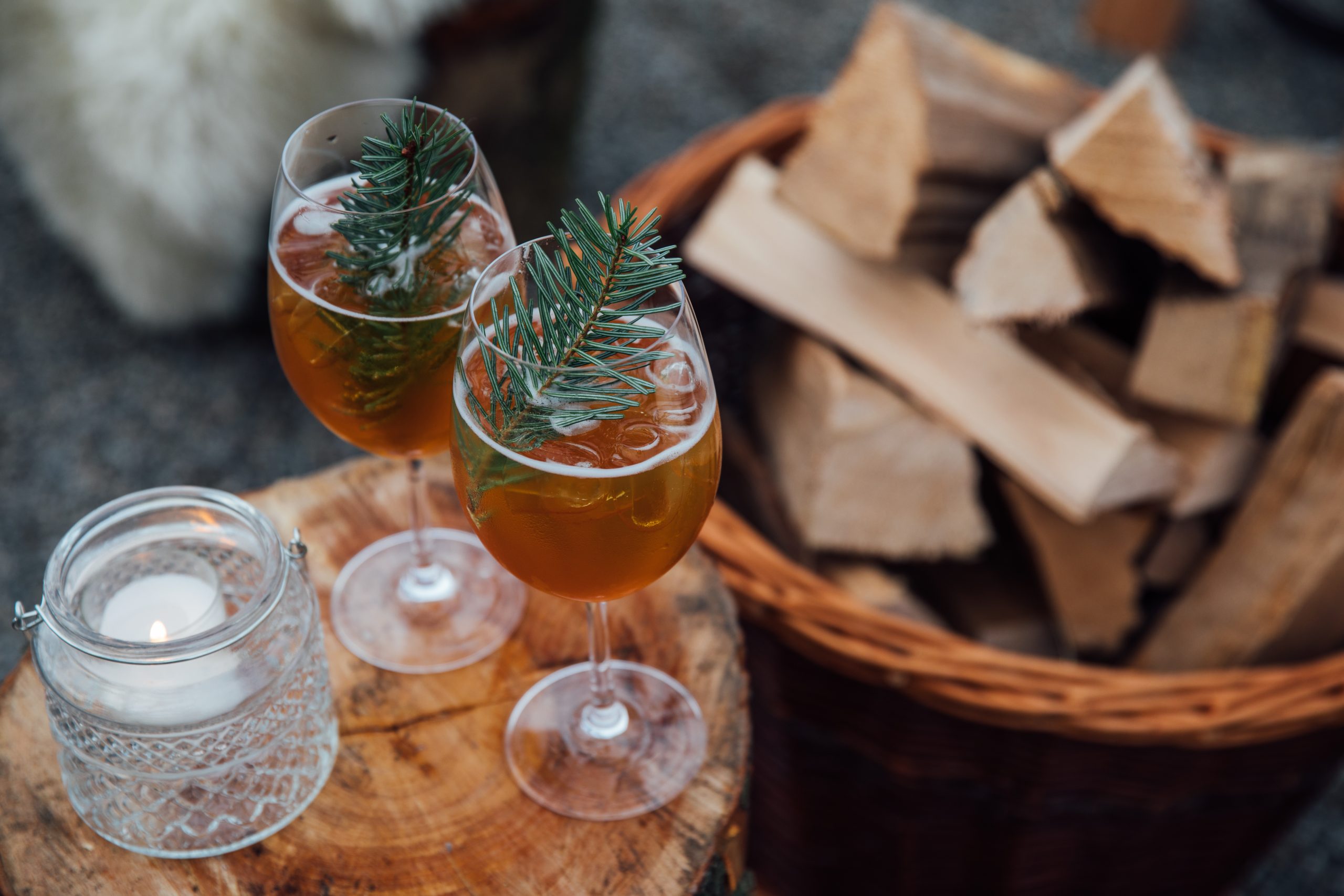 Zwei Gläser mit bernsteinfarbenen Getränken, die mit Tannenzweigen garniert sind, stehen im Chalet am See auf einer hölzernen Fläche, daneben eine leuchtende Kerze in einem Glashalter und ein Korb mit Brennholz im Hintergrund.