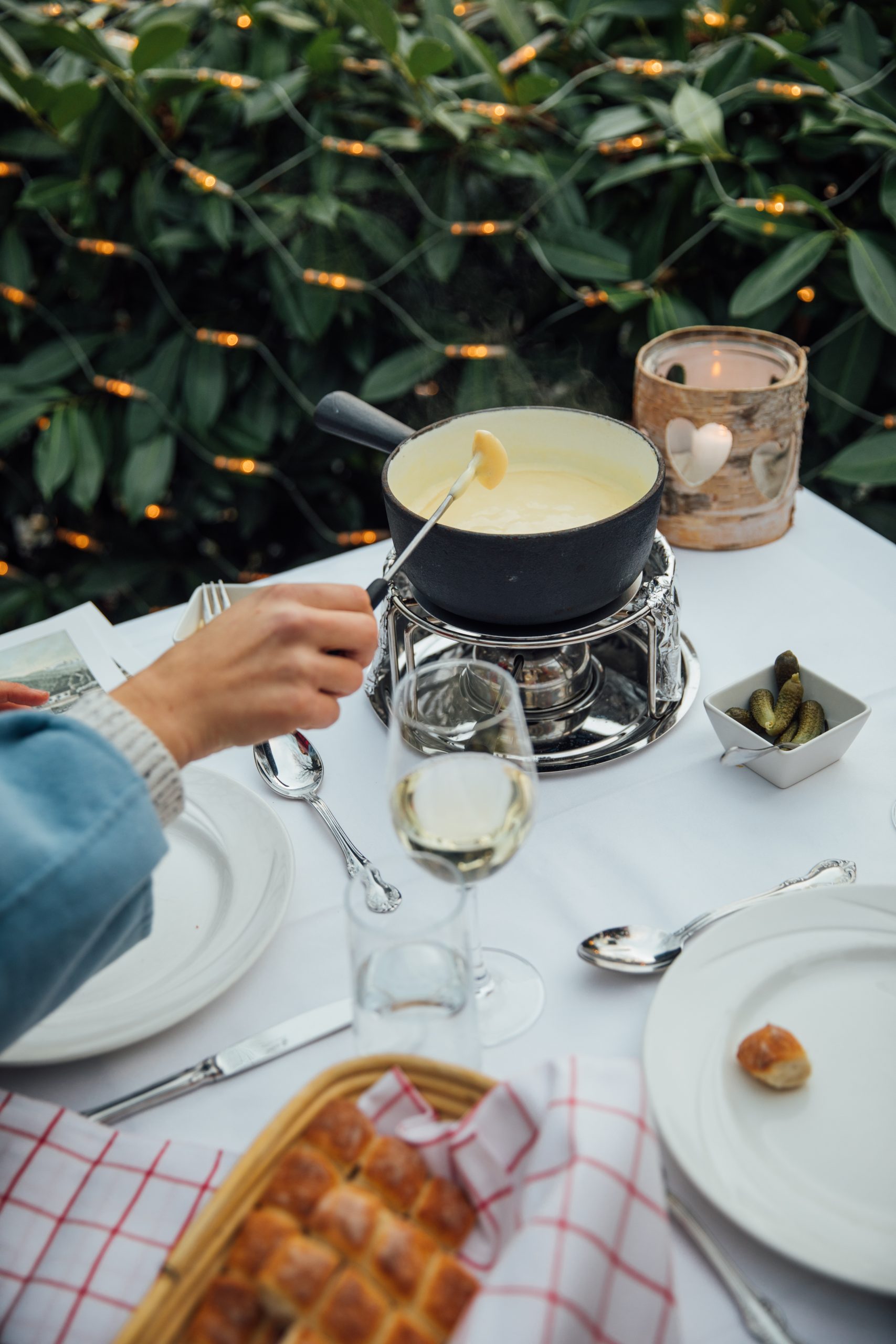 Eine Person taucht Brot in einen Topf mit Käsefondue am Tisch im Freien des Restaurants Seegarten, der mit Tellern, Besteck, Wein, Essiggurken und einer Kerze gedeckt ist, umgeben von Grünzeug und Lichterketten.