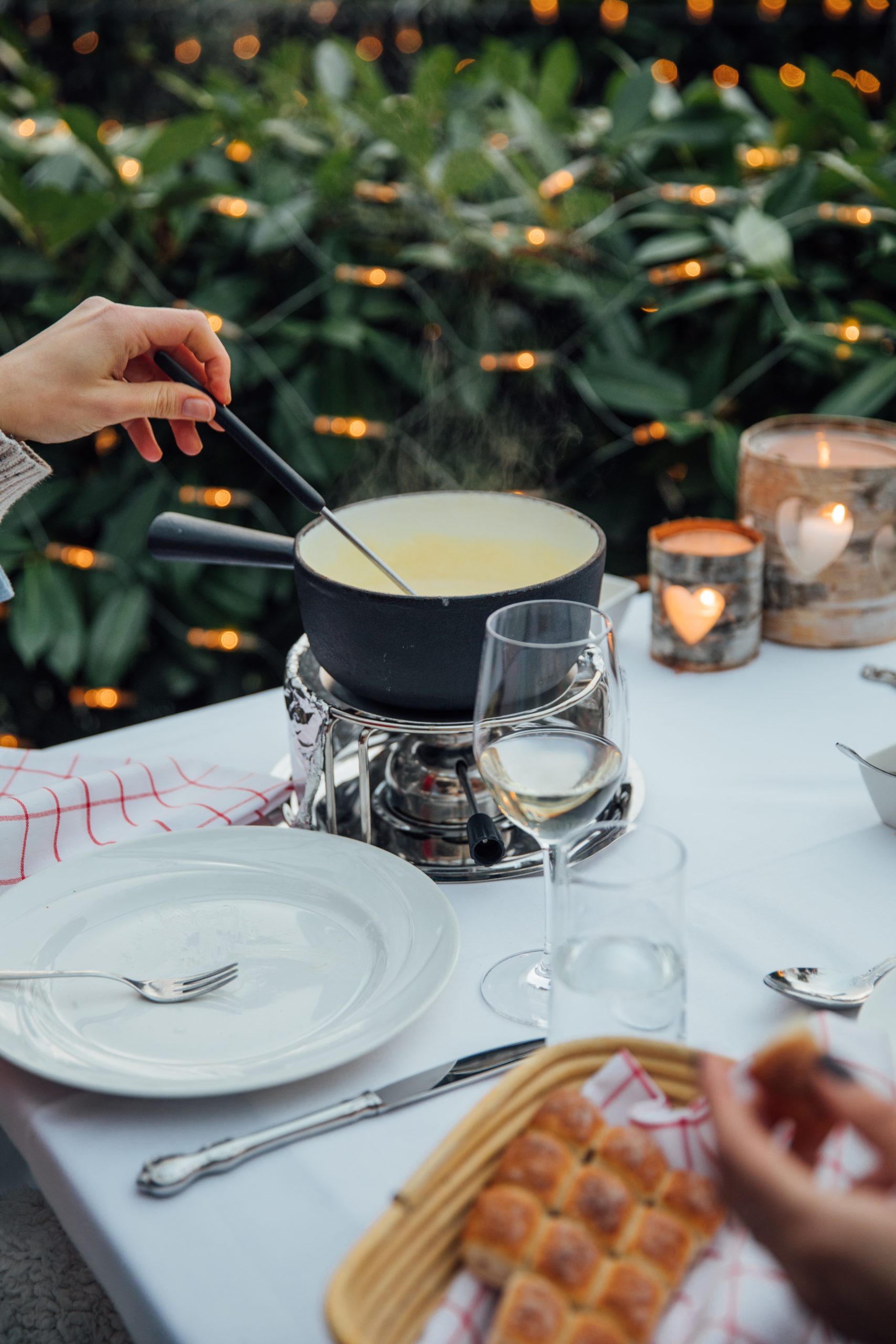 A person dips a fork into a pot of cheese fondue on a white-clothed table set with plates, wine glasses, bread, and candles at a chalet by the lake, with greenery and string lights in the background.